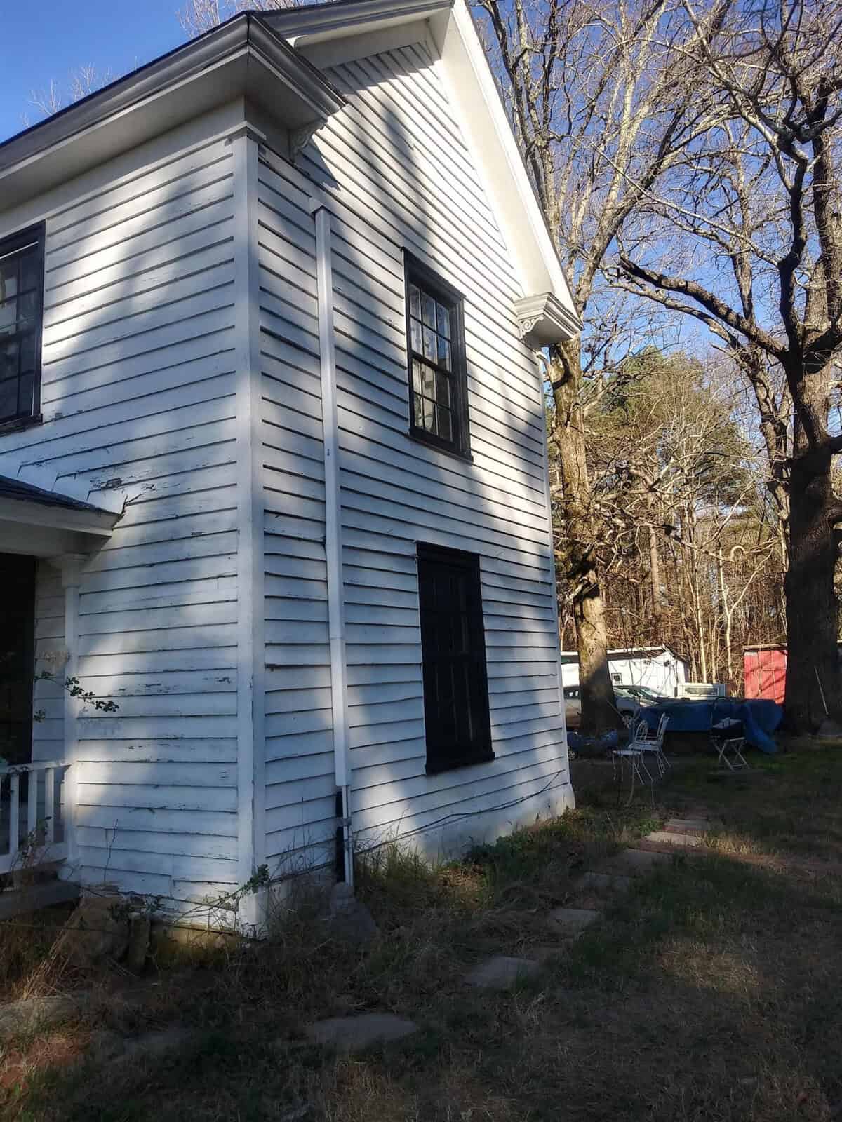 A white wooden house with black shutters on a sunny day, partially casting shadows on the wall, with an overgrown grassy front yard and a collection of blue chairs to the side.