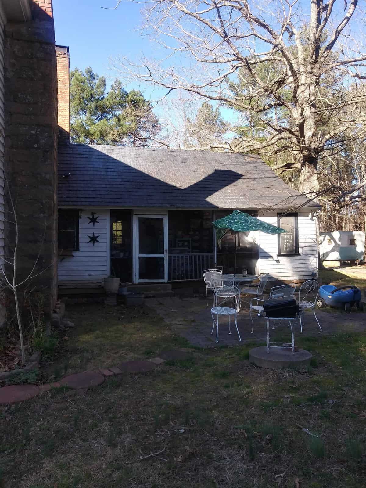 A single-story house with a porch and outdoor seating area surrounded by trees and a lawn, with various items scattered around the yard.