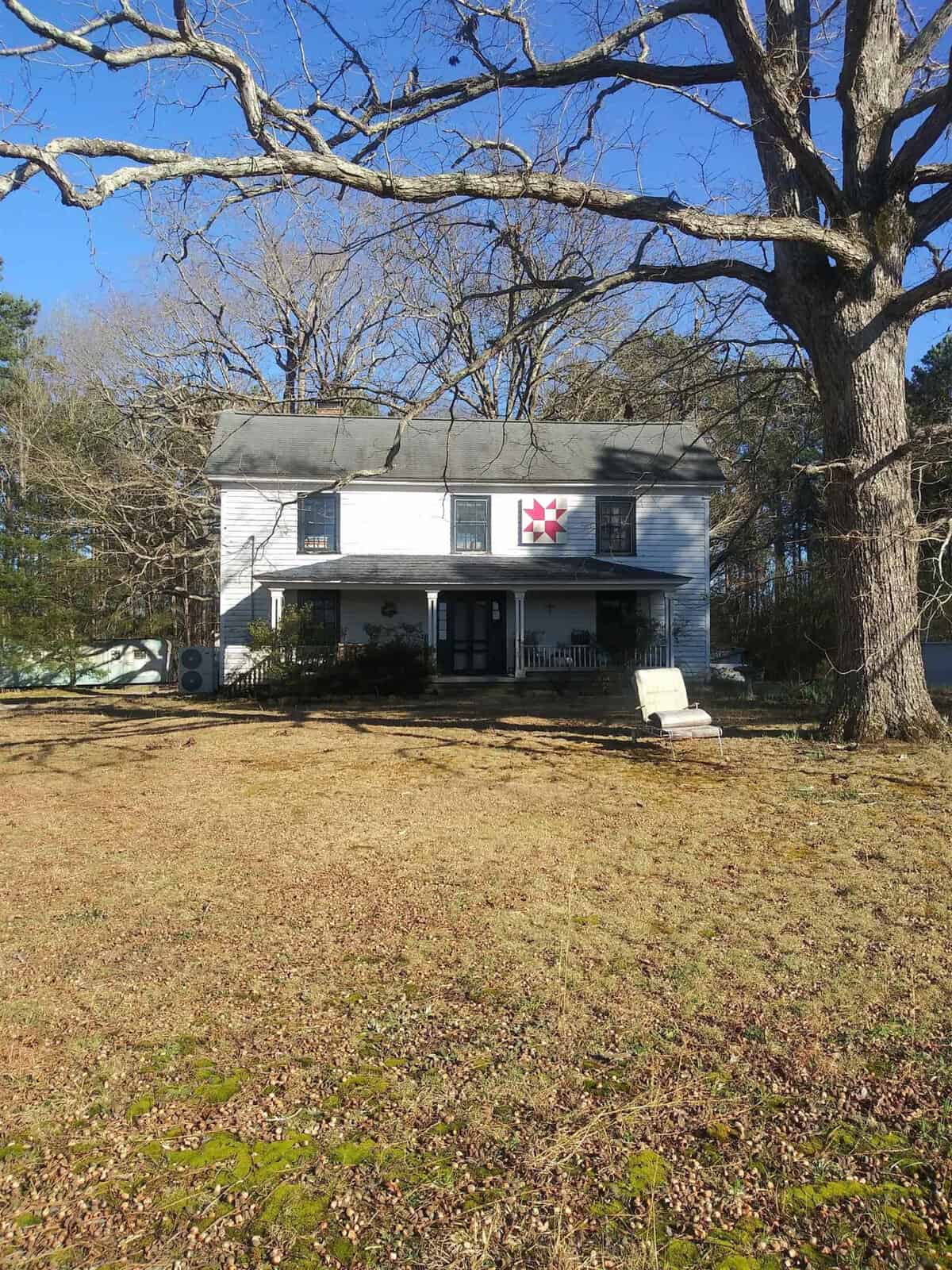 A two-story house with white siding under a clear sky, featuring a red star decoration on the front, surrounded by leafless trees and a lawn with patchy grass.