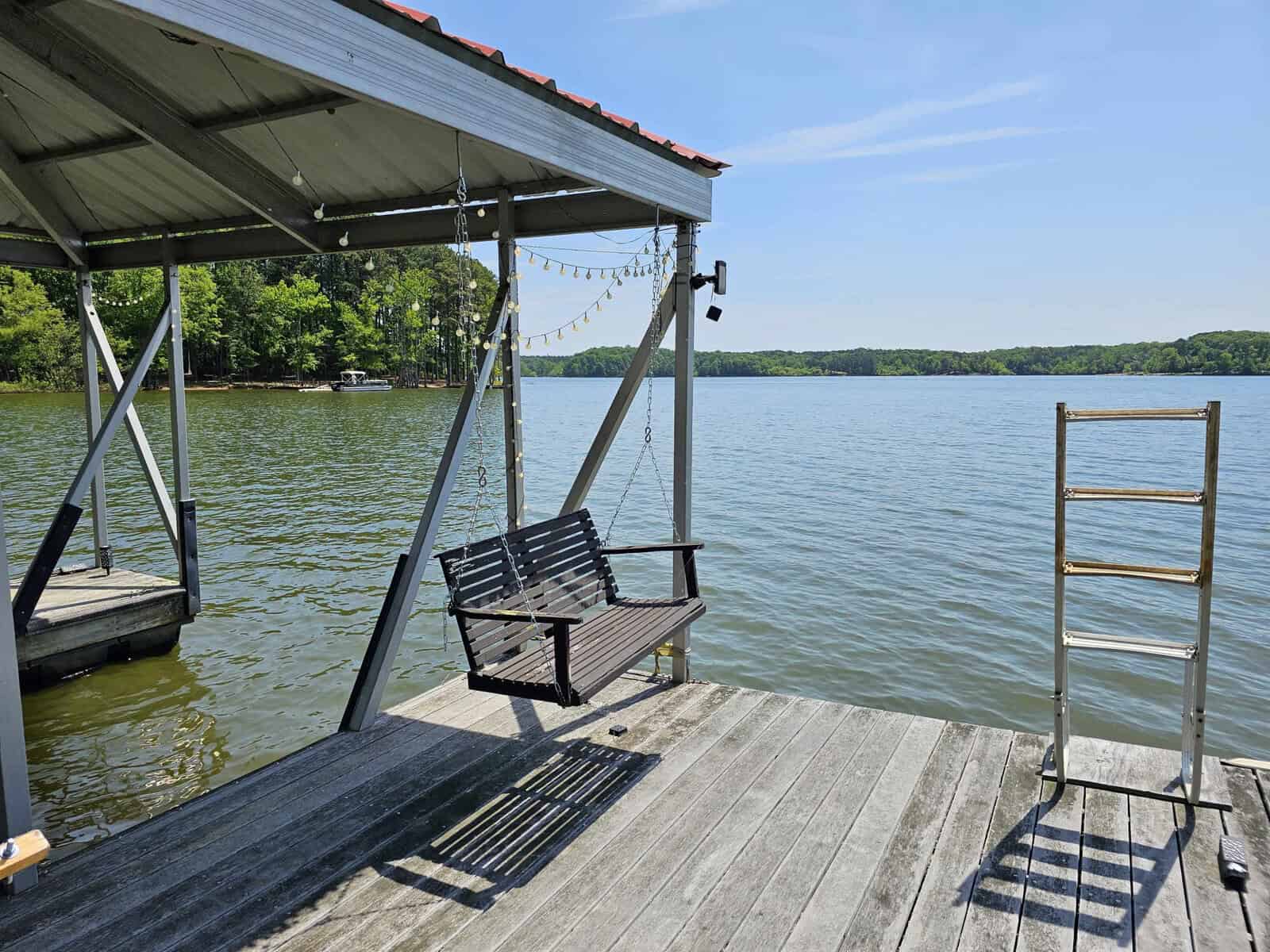 A wooden dock with a swinging bench under a roof, overlooking a calm lake on a clear day at 306 Waters Edge Loop, Henderson NC.