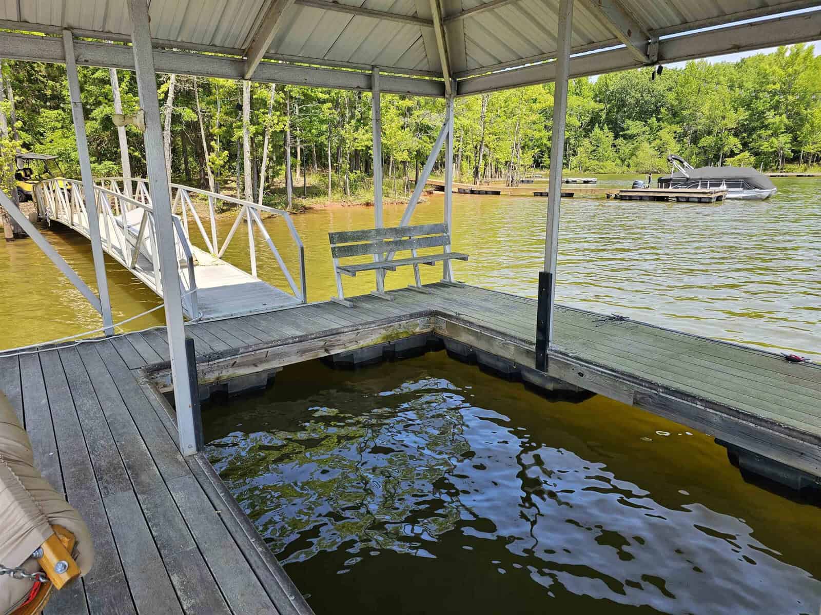 A wooden dock with a metal roof extends over a calm body of water, featuring a bench and a walkway leading back to land, with a pontoon boat moored nearby.