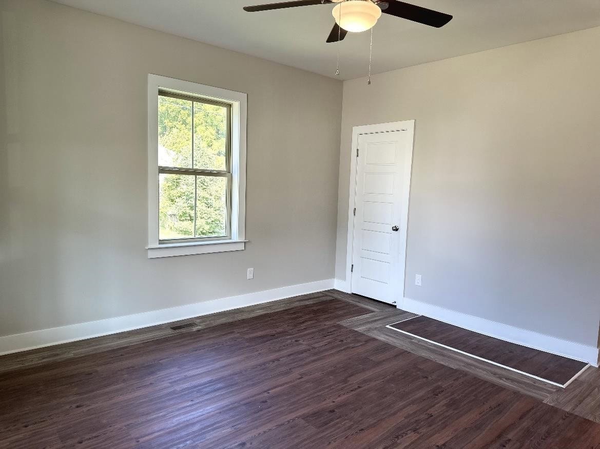 An empty room with light gray walls, dark wooden flooring, a closed white door, and a window with a view of greenery outside. there's a ceiling fan with lights installed in the center of the room.