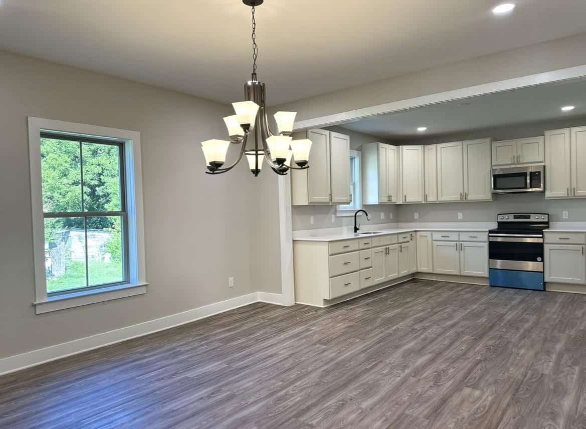A modern kitchen with white cabinets, stainless steel appliances, and a hanging light fixture, with a view of greenery outside the window.