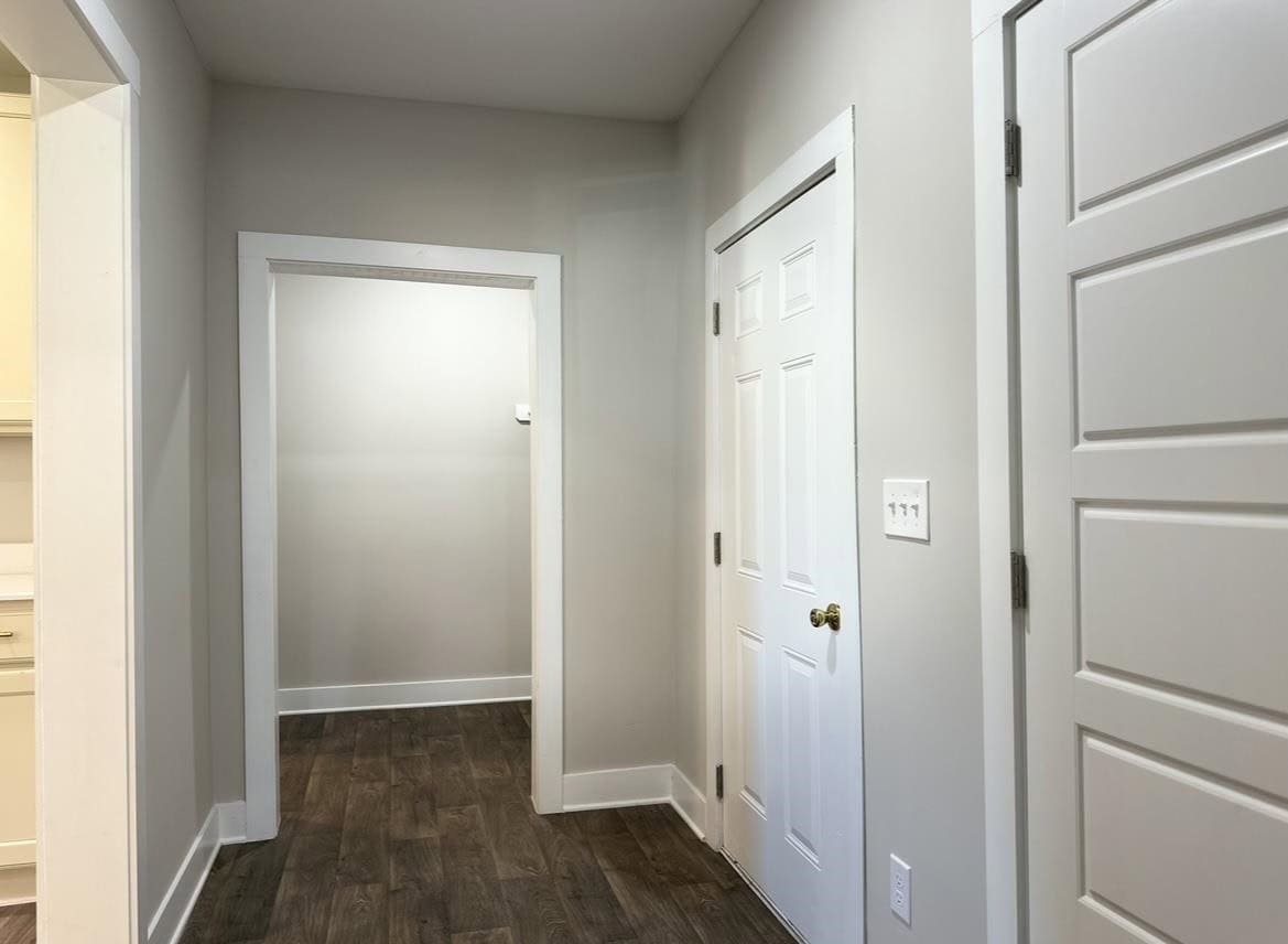 An empty hallway featuring a wooden floor, with two closed white doors and an open doorway leading to another room; a light switch and electrical outlet are visible on the right wall.