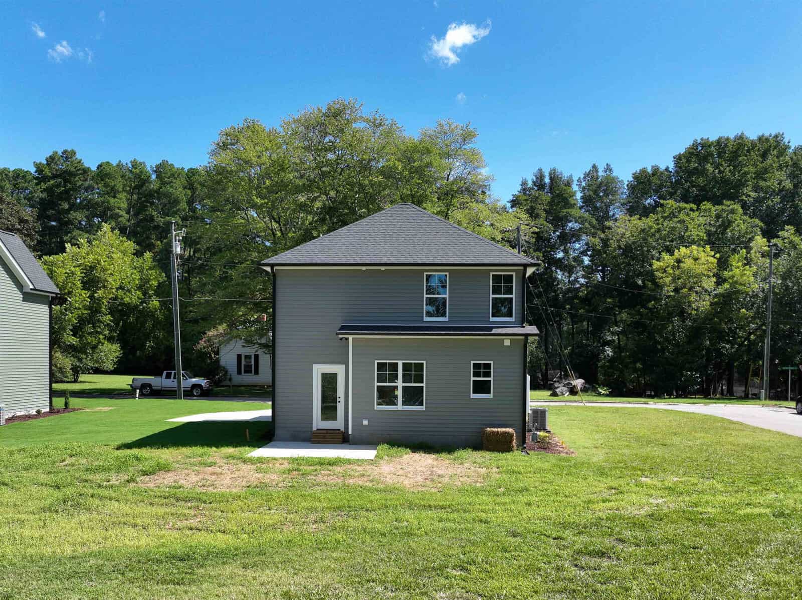A two-story gray house with a gable roof is situated on a grassy lawn. A small concrete path leads to the front door. Trees and an adjacent house are visible in the background under a clear blue sky.