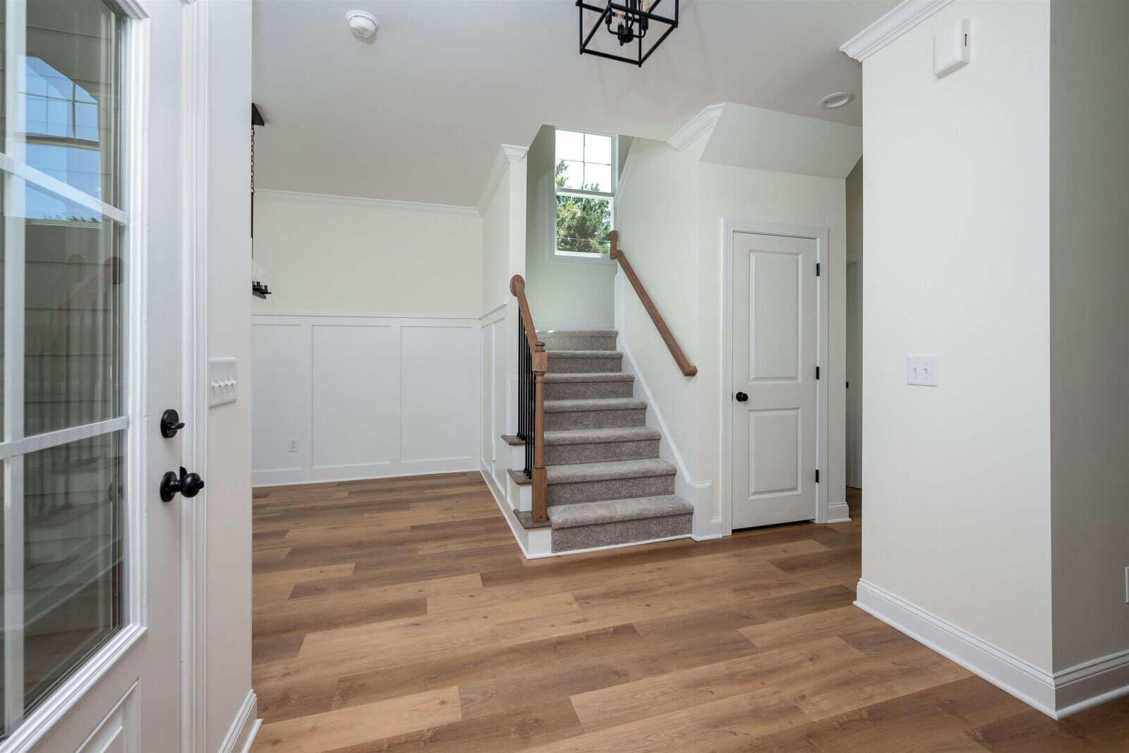 A bright entryway featuring wooden flooring and white walls. Stairs with a wooden banister lead upwards. A white door with a black handle is on the right, and a glass-paneled door is on the left. A ceiling light fixture hangs above.