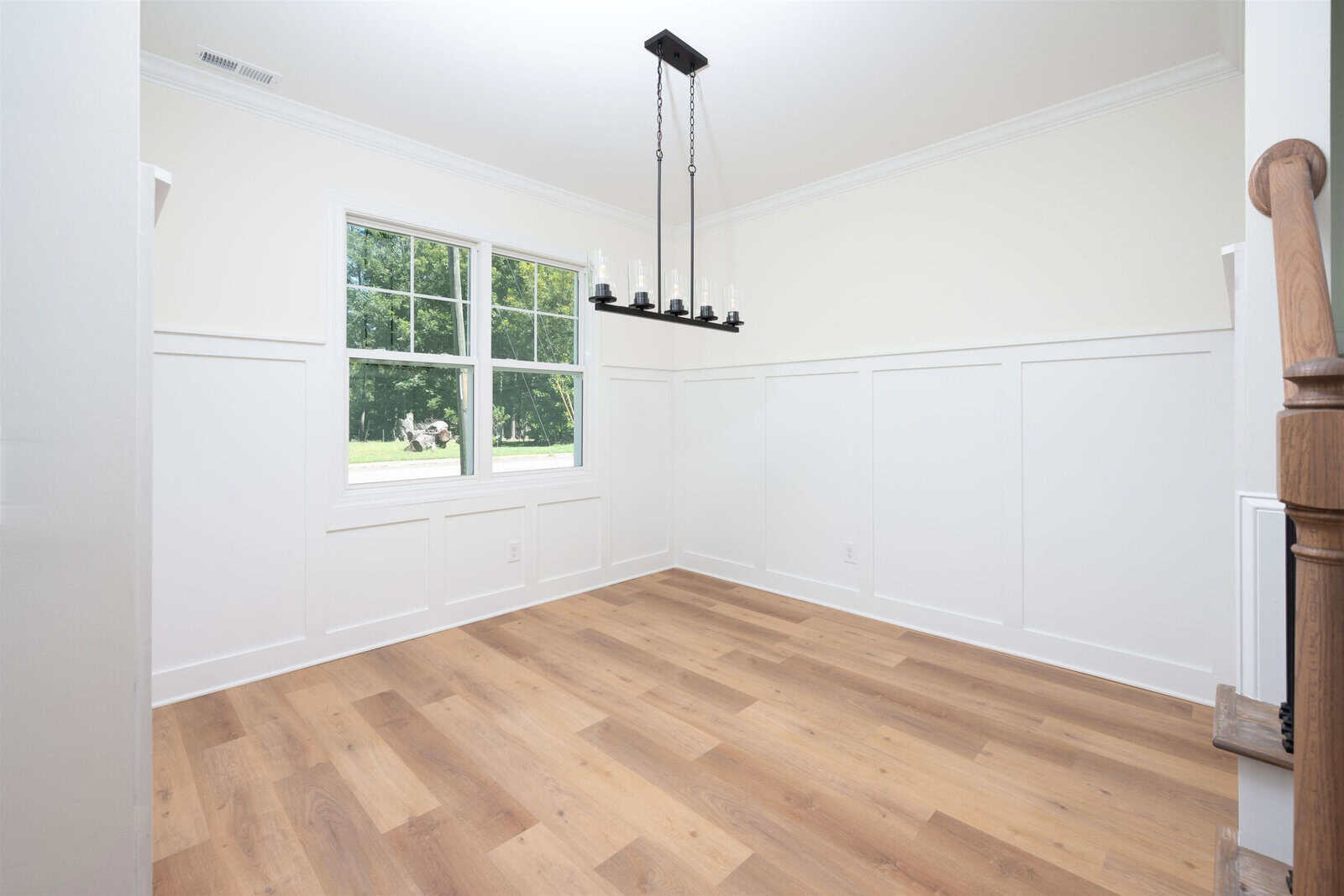 A bright, empty room with white walls and a light wood floor. A large window with a view of greenery is on the left wall. A black chandelier with candle-shaped bulbs hangs from the ceiling. Wainscoting decorates the lower walls.