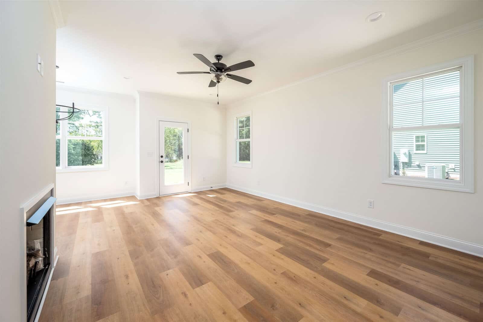 A bright, empty living room with light hardwood floors and pale walls. There's a ceiling fan and several windows. A door with a window leads outside, allowing natural light to fill the space.