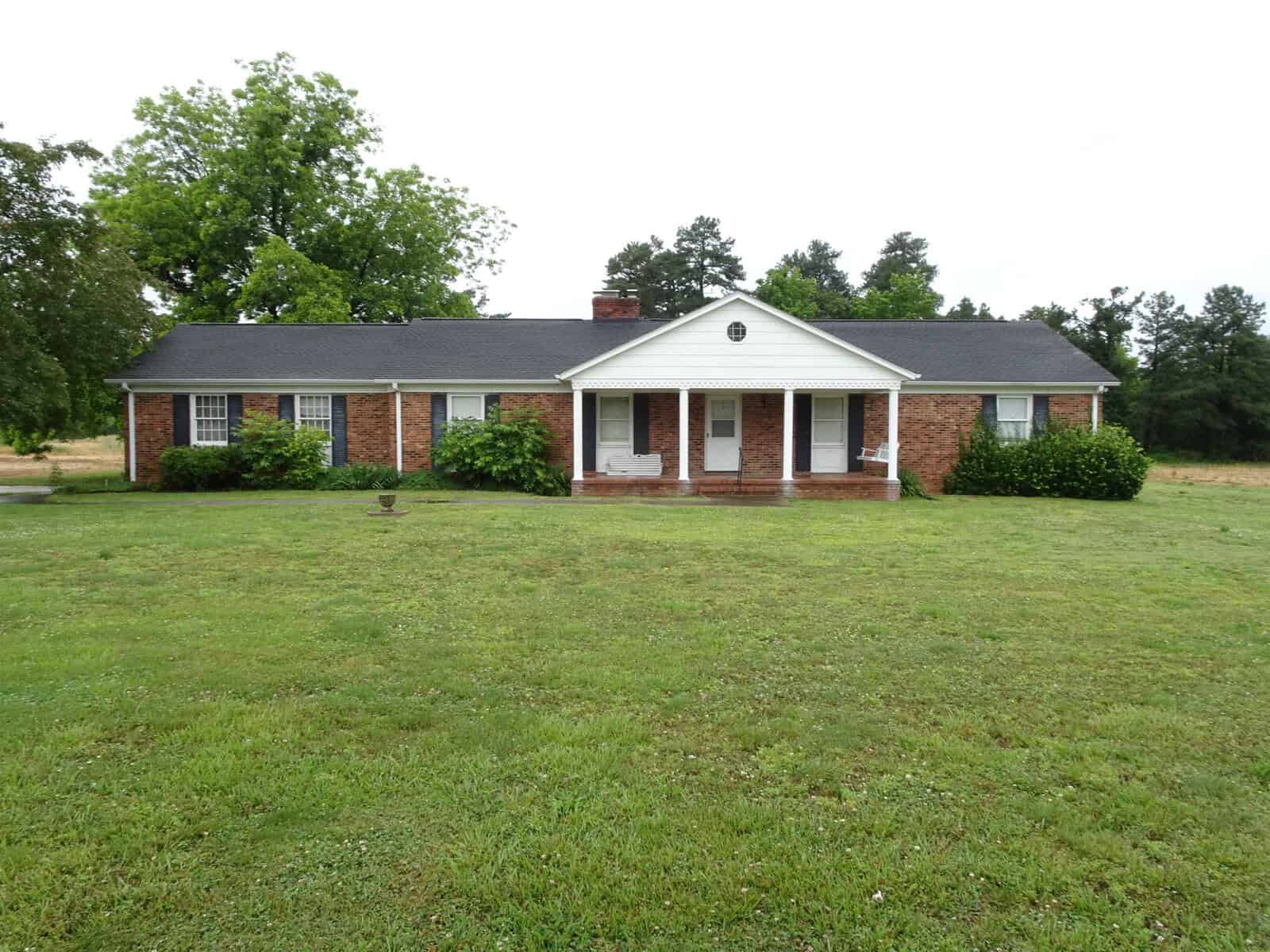 A single-story brick house with white trim and a central entryway, set back from a large front lawn with scattered trees and shrubs.