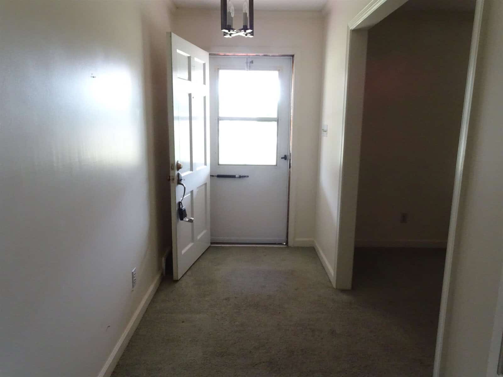 An empty hallway with carpeted flooring leading to a closed white door with a window, flanked by a small table to the left and a light fixture overhead.