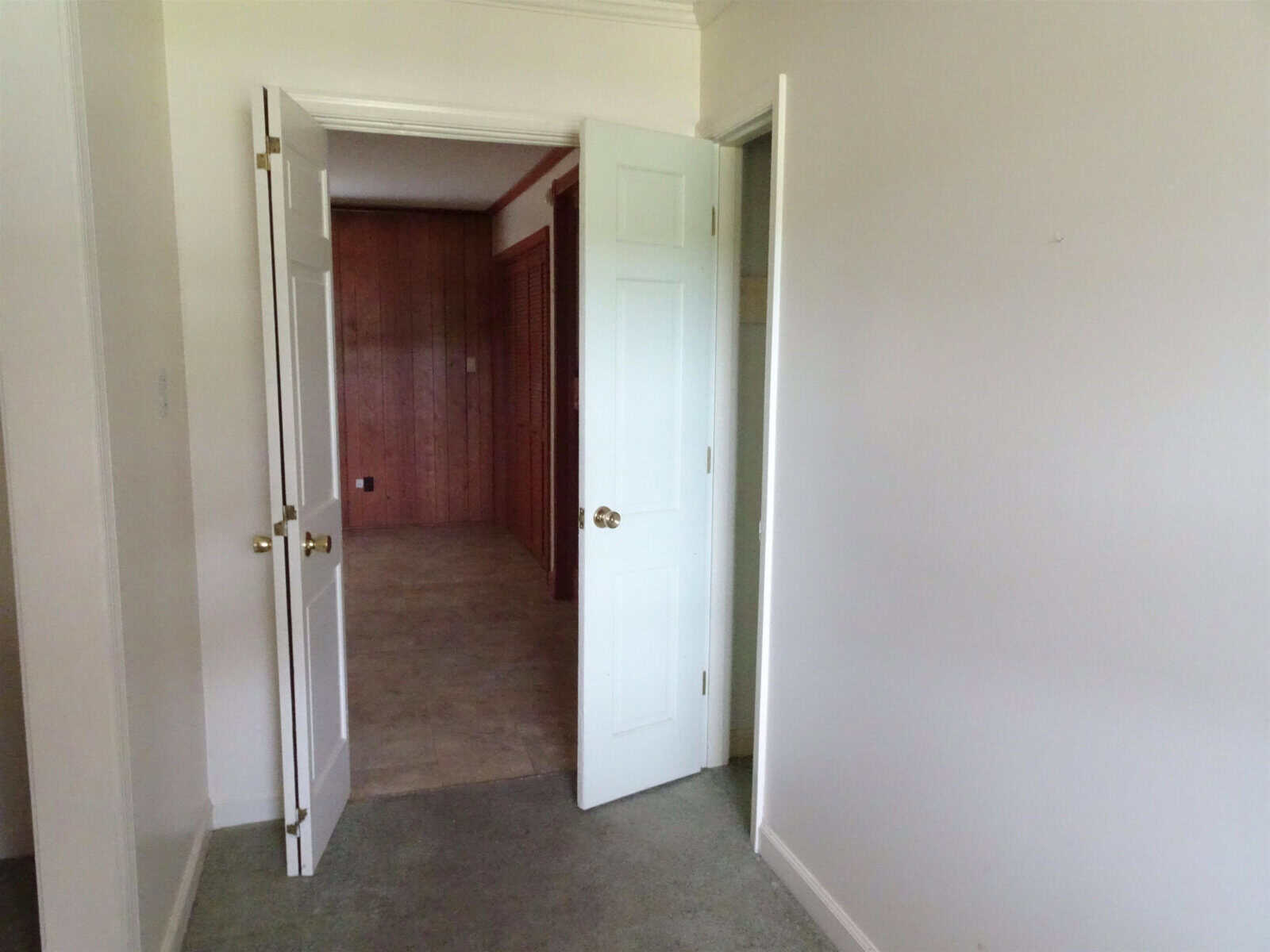 An empty room with an open door leading to another room with wood-paneled walls and carpeted floors.
