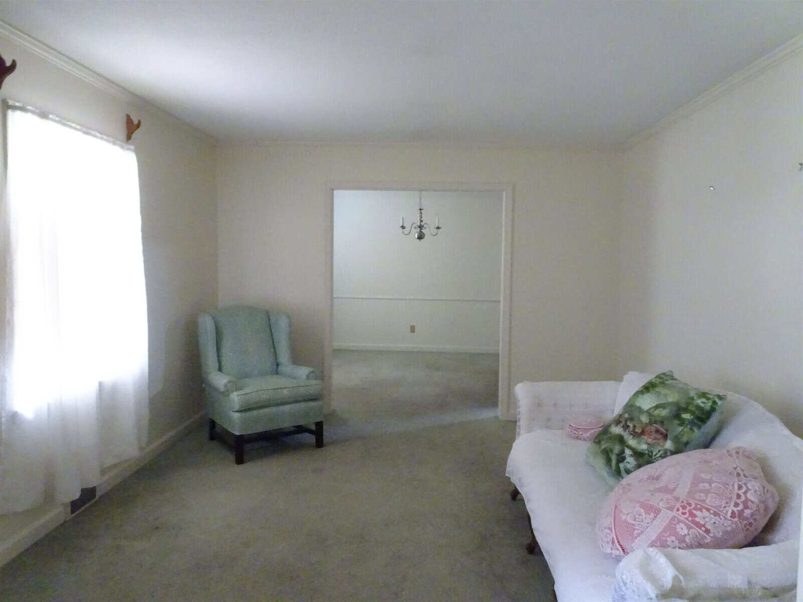 A sparsely furnished room with an armchair, a sofa with decorative pillows, a chandelier, and a closed curtain on one window.