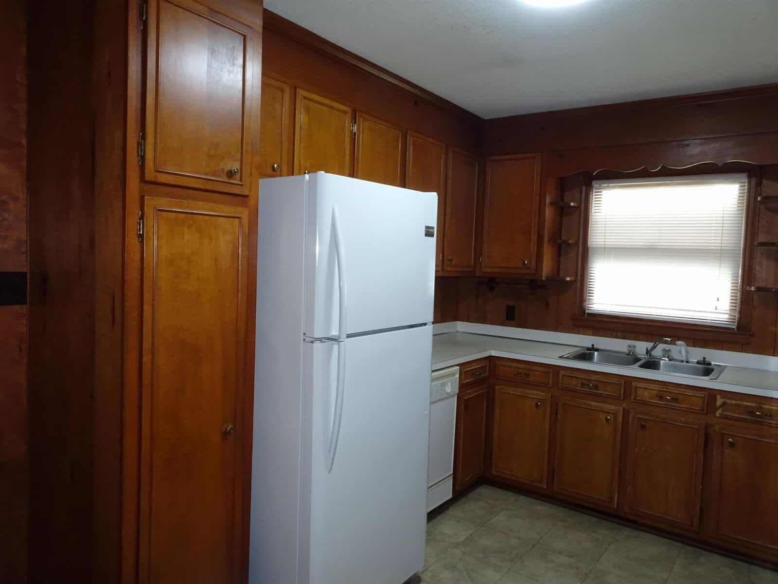 A residential kitchen featuring wooden cabinetry, a white refrigerator, a sink beneath a window with a closed blind, and light-colored countertops.