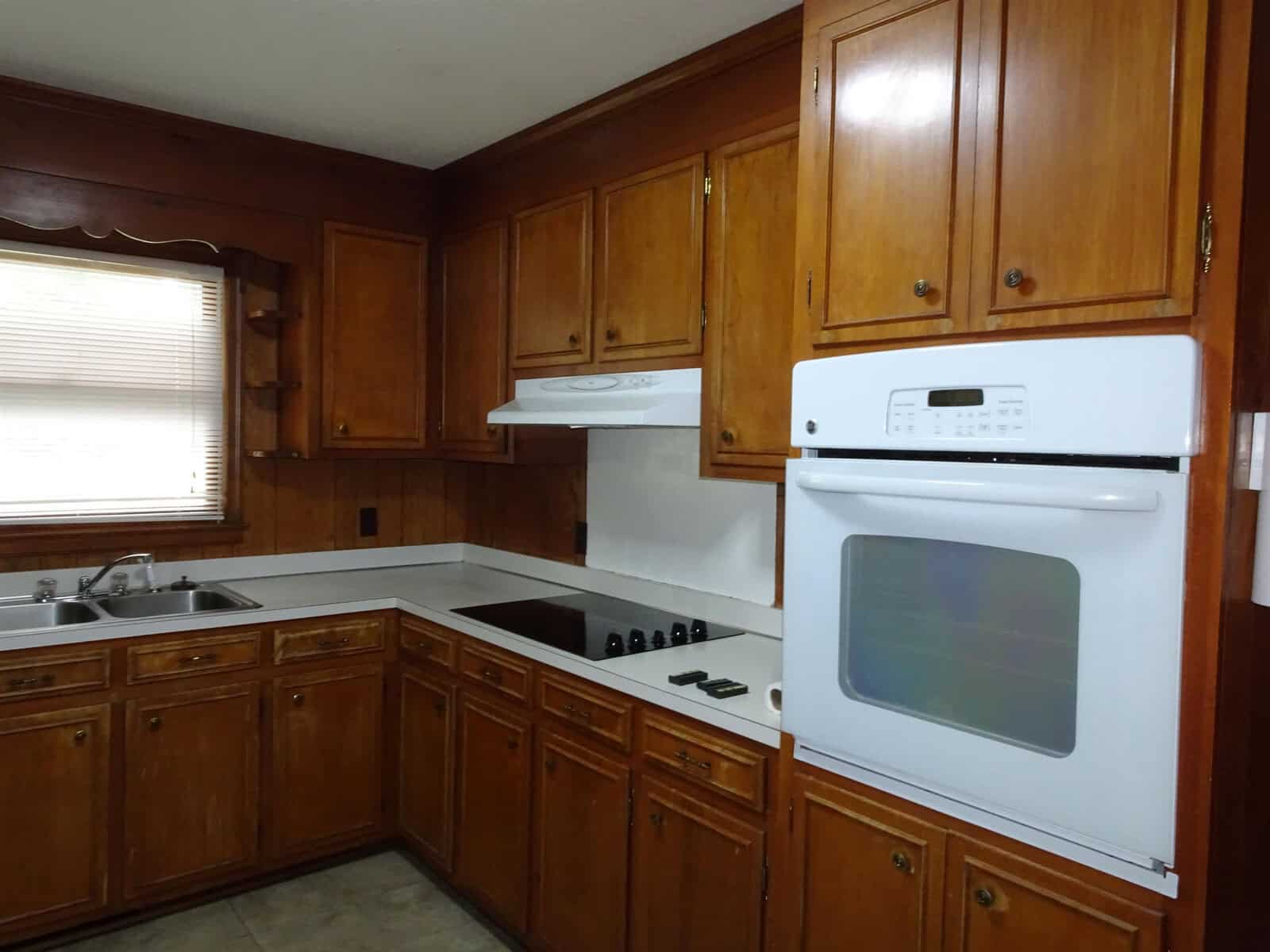 A traditional kitchen featuring wooden cabinets, white appliances including a stove and hood range, with a countertop and a double basin sink under a window with horizontal blinds.