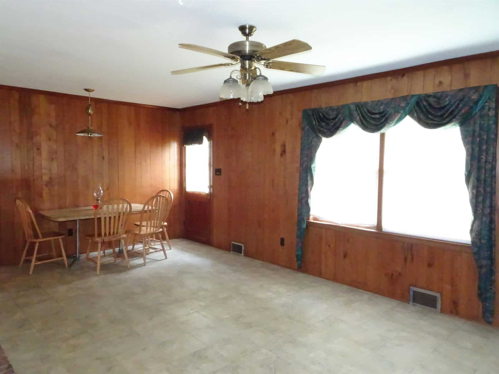 A spacious room with wood paneling on the walls, a ceiling fan, and a dining area with a wooden table and chairs near a large window with draped curtains.