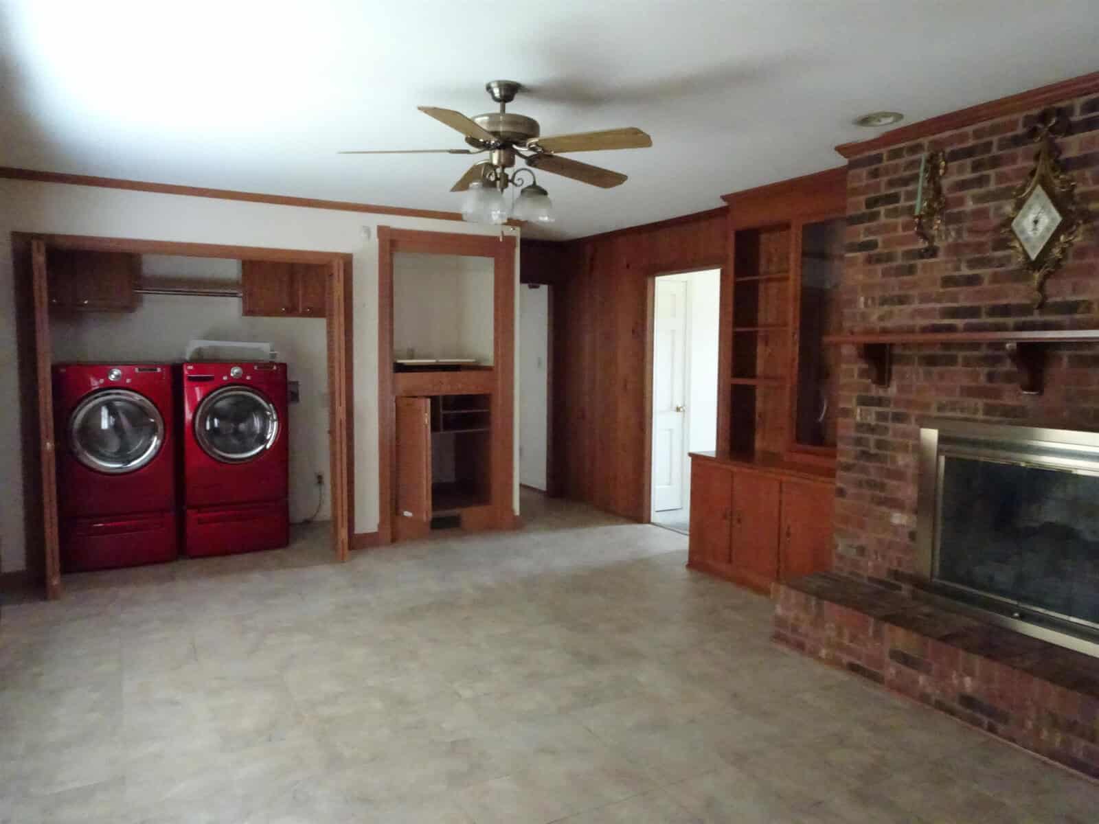 A spacious room with a laundry area on the left, featuring a red washer and dryer, and a brick fireplace on the right. there's an empty built-in shelving unit near the laundry area, and a ceiling fan is installed in the center of the room. the floor is tiled, and there's a white door at the back.