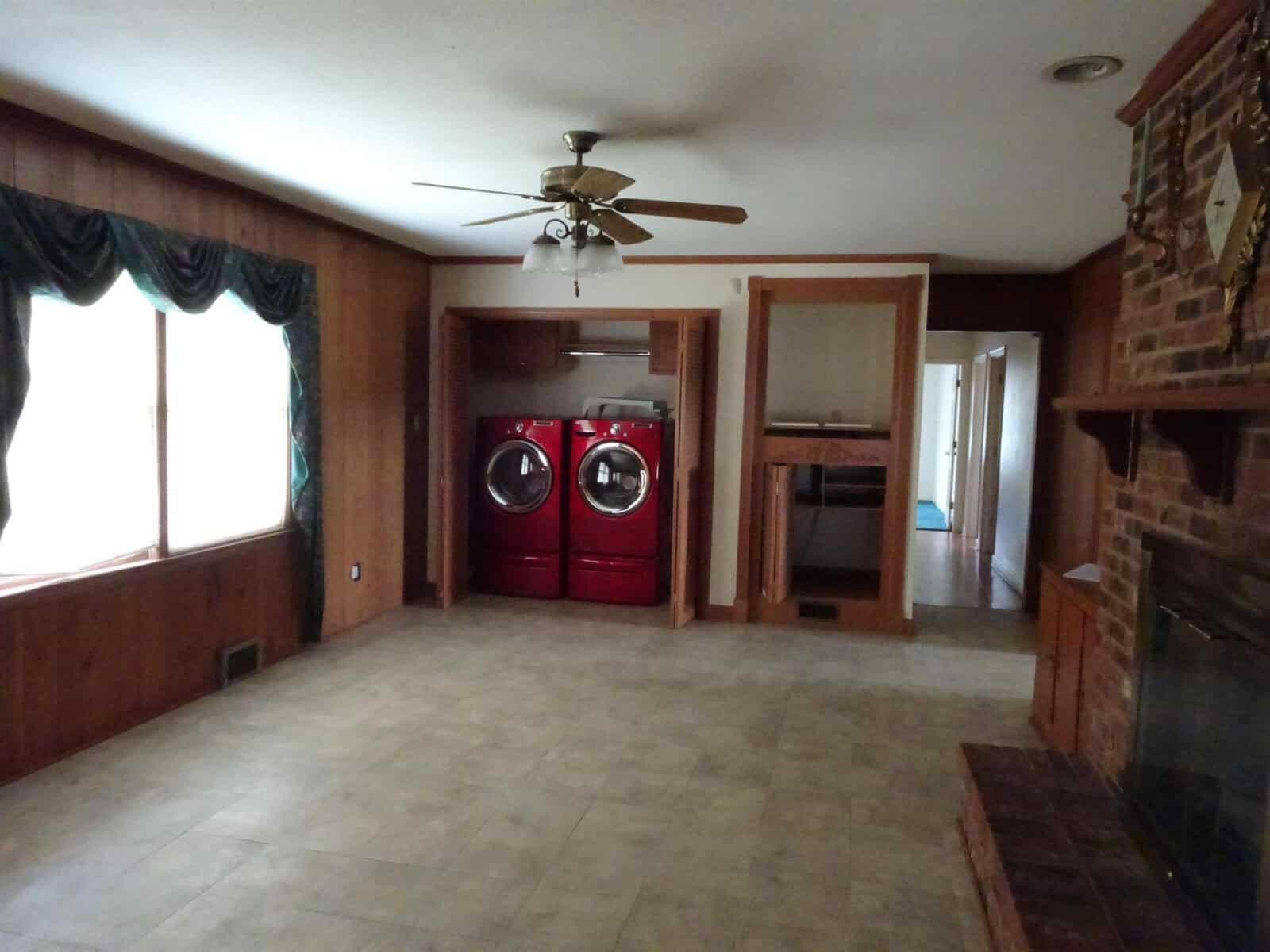 A spacious room featuring a tile floor with a ceiling fan, dark wood paneling on the walls, a fireplace on the right, a pair of red washing appliances by the back wall, and a large window with dark drapes on the left.