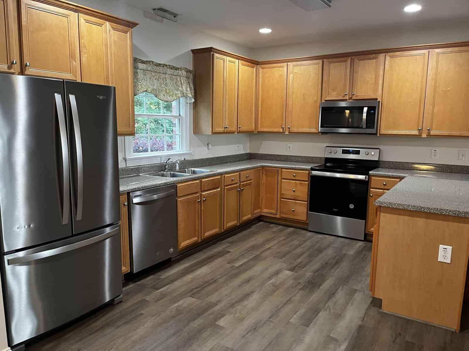 A modern kitchen with wooden cabinets, granite countertops, stainless steel refrigerator, and black appliances including a dishwasher, oven, and microwave.