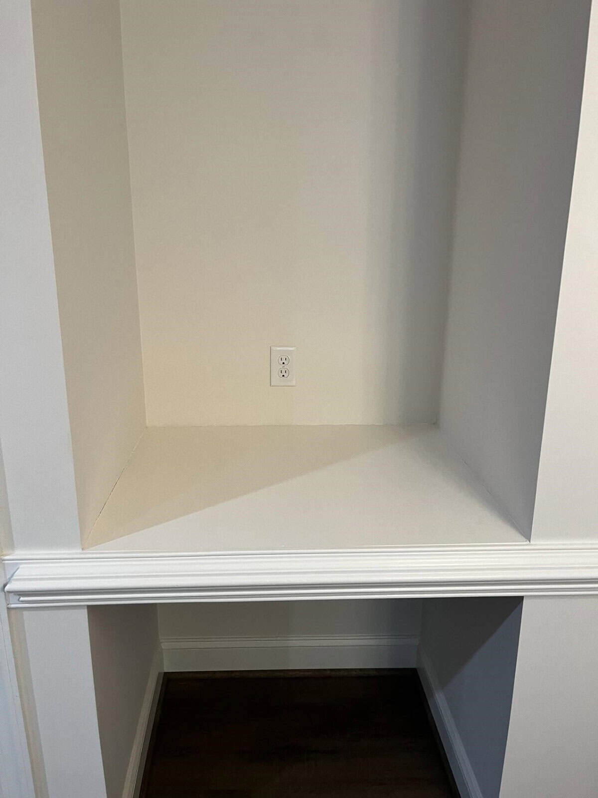 An empty nook with white walls and a single electrical outlet, above a white baseboard and wooden flooring.