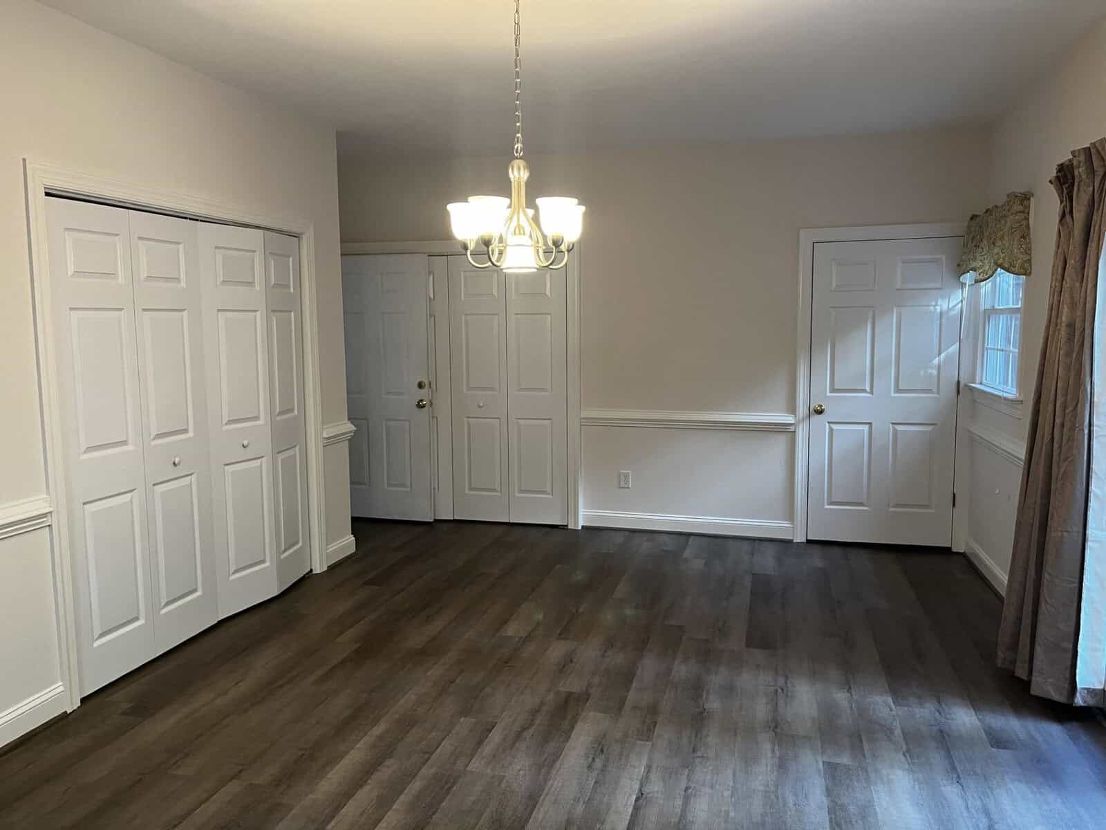 A well-lit, empty room featuring dark wood flooring, white walls, a white chair rail, a pendant light fixture, two closed white doors, and one partially open white closet door, with a glimpse of a window treatment on the right side.