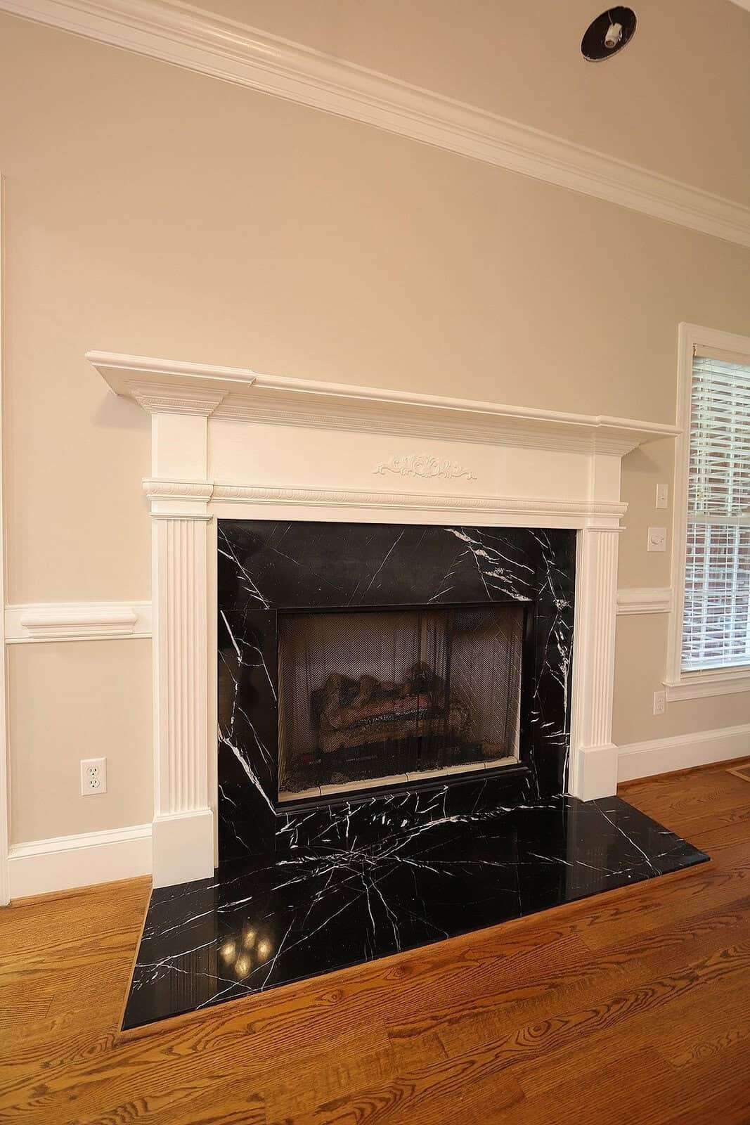 A traditional style white mantelpiece with a marble surround and hearth, enclosing a gas fireplace, set against a neutral wall with white wainscoting, adjacent to a window with white blinds in a residential home.