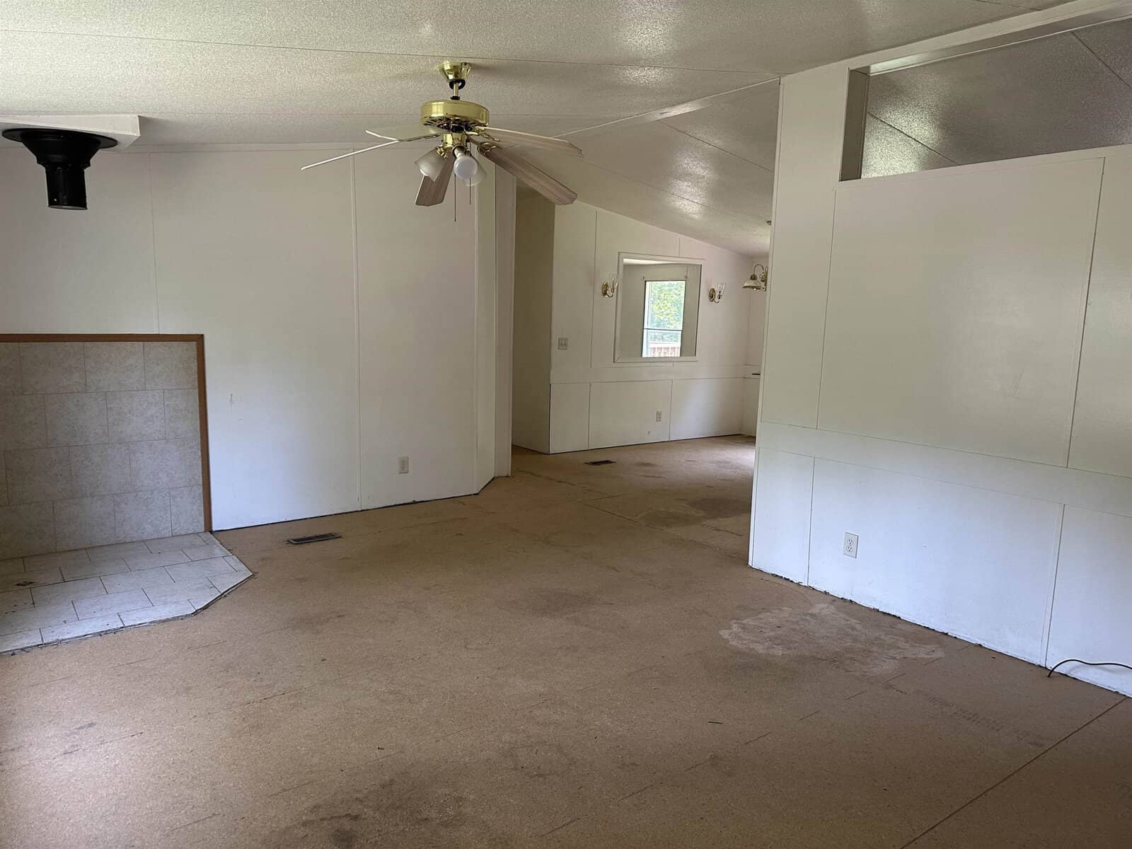 An empty room with beige carpeting showing signs of wear and patches of missing carpet, white walls, a ceiling fan, and a fireplace hearth on the left side.