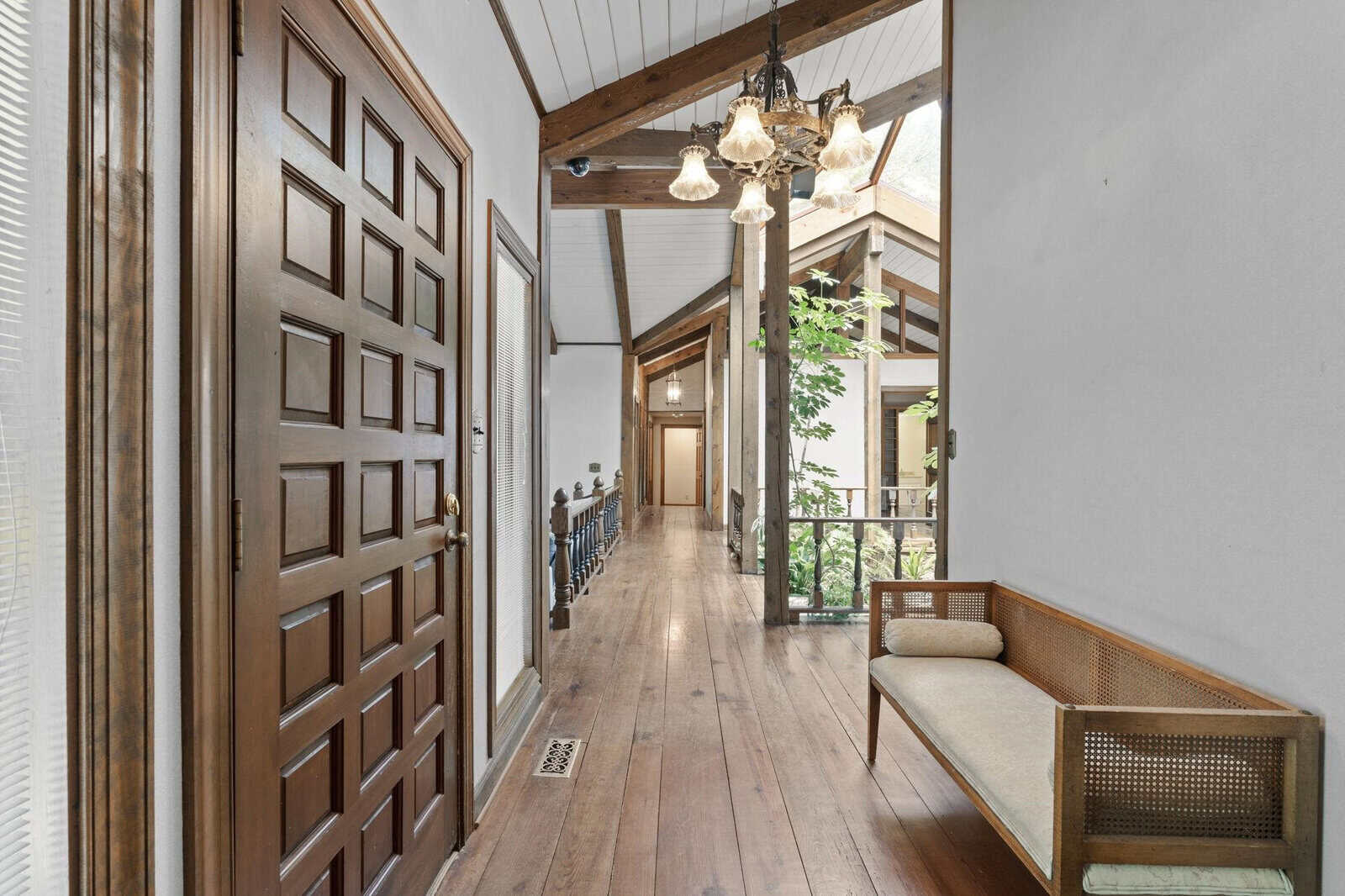 A spacious interior hallway featuring wooden floors and walls, with a bench to the right and a glass-paneled door open to the next room, illuminated by natural light and a hanging chandelier.