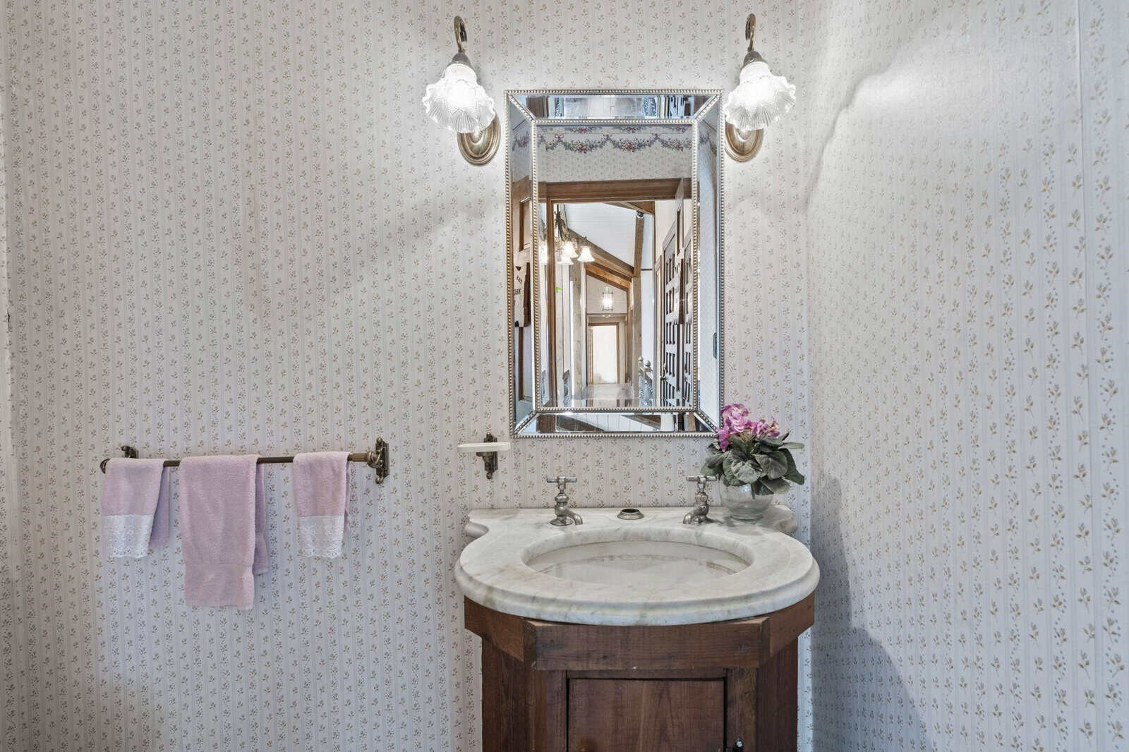 A traditional bathroom interior with a wooden vanity cabinet, a white basin, a silver mirror, and wall-mounted lamps, with floral wallpaper in the background.