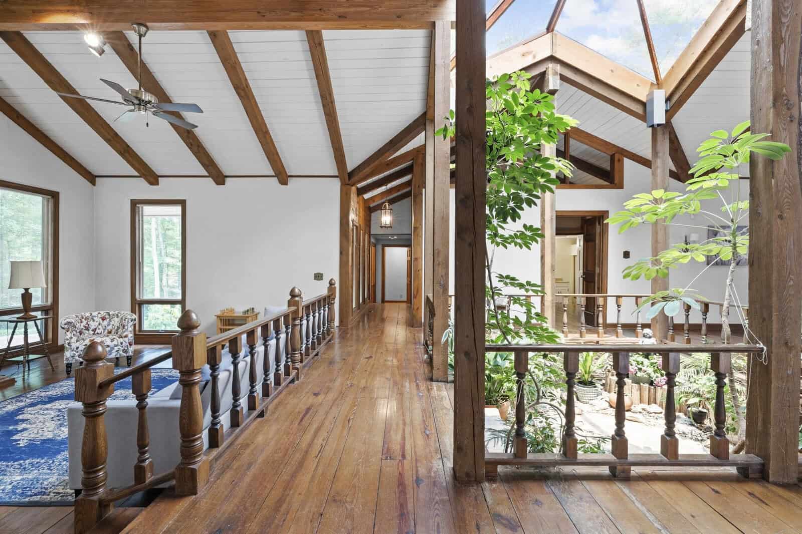 An interior wooden walkway with a spindle railing overlooks a sunlit atrium with potted plants, leading from a room with ceiling fans to another part of the house.