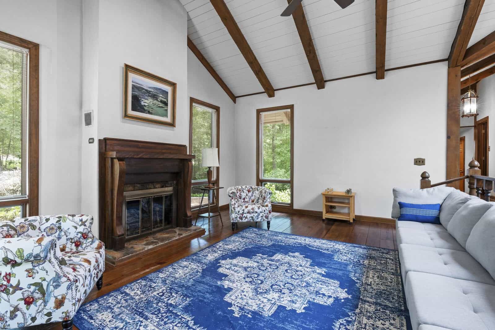 A spacious living room with a vaulted ceiling featuring exposed wooden beams, white walls, a dark wood fireplace, and furnished with patterned chairs, a white sofa, and a large blue area rug.