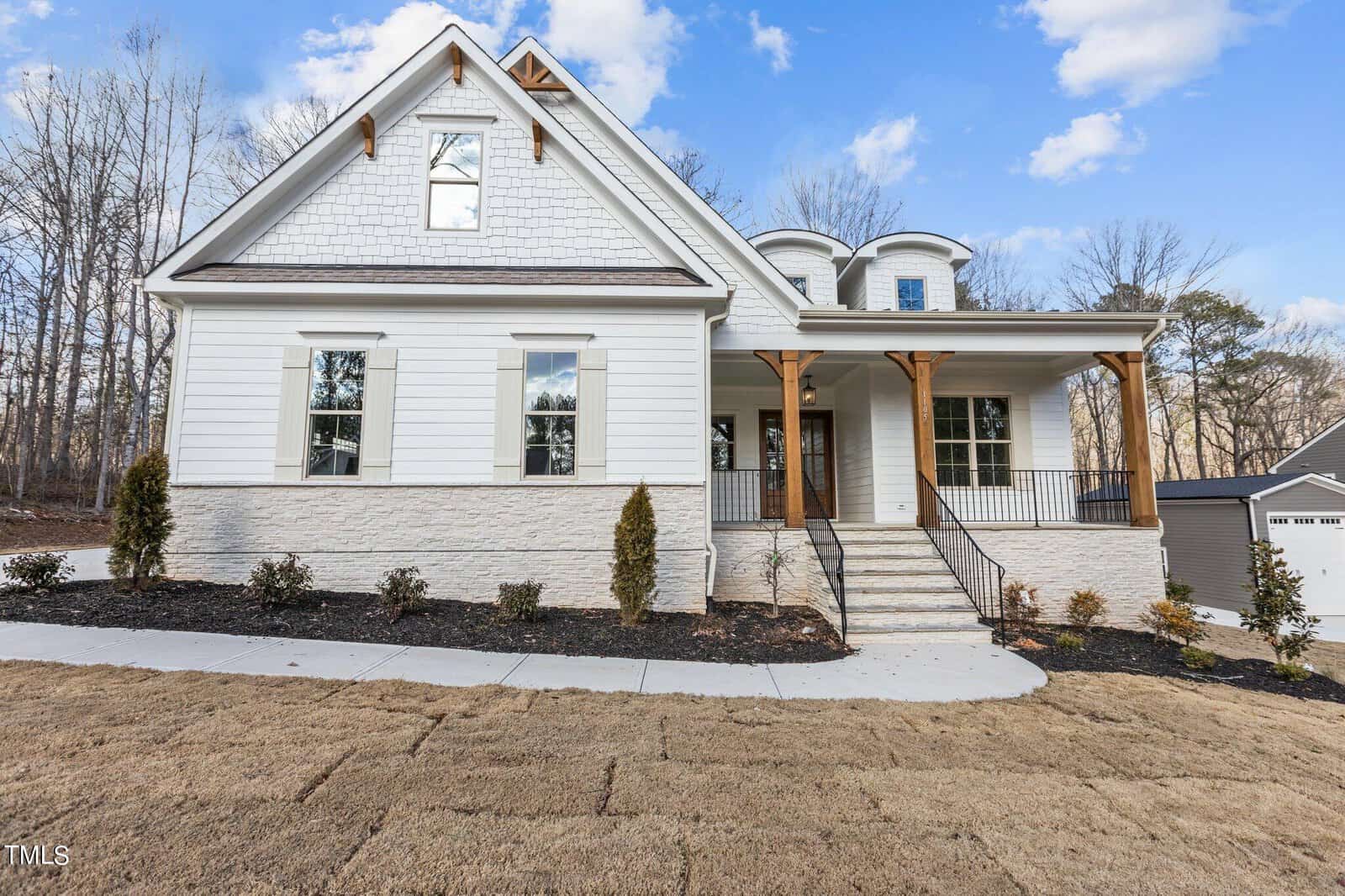 A white two-story house with a gabled roof and wooden pillars on the front porch. The house has multiple windows and a stone foundation. The lawn is newly planted and bordered by young shrubs. Trees are visible in the background.