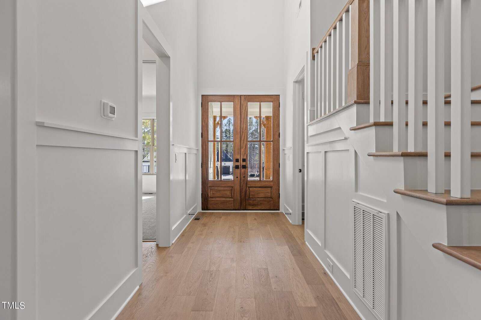 A bright and spacious hallway with wooden flooring leads to double wooden doors with glass panels. Walls are painted white, and a staircase with white railings and wooden steps is visible on the right. There is a room entrance on the left.