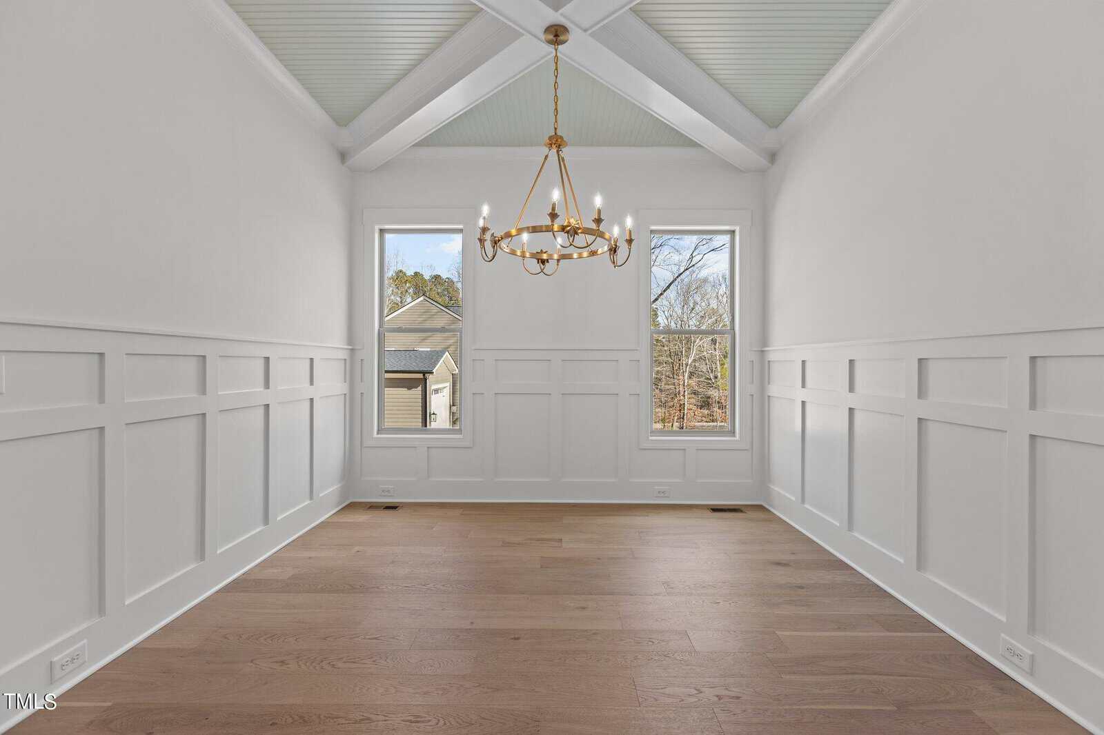 A bright, empty room with white walls and wainscoting features two tall windows and a ceiling with wooden beams. A gold chandelier hangs from the ceiling, and the floor is made of light brown wood. Trees are visible outside the windows.