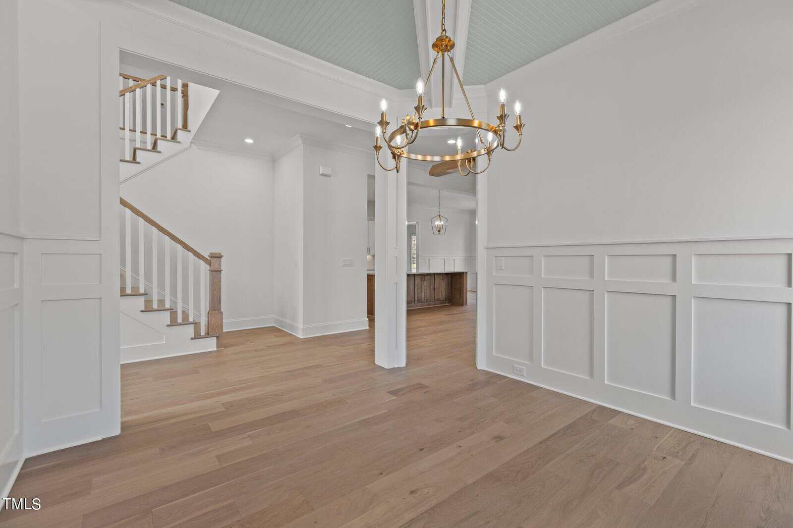 A spacious interior features a room with light wood flooring and white wainscoting. A gold chandelier hangs from the ceiling. In the background, a staircase with wooden handrails and a kitchen area are visible. Walls are painted white.