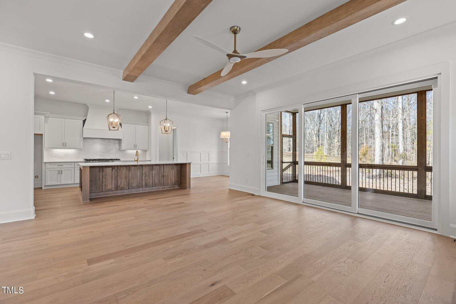 A spacious, modern interior with light wood flooring and a ceiling fan. The open kitchen features an island with pendant lights. Large sliding doors lead to a screened porch with views of a wooded area. White walls and wooden ceiling beams complete the space.