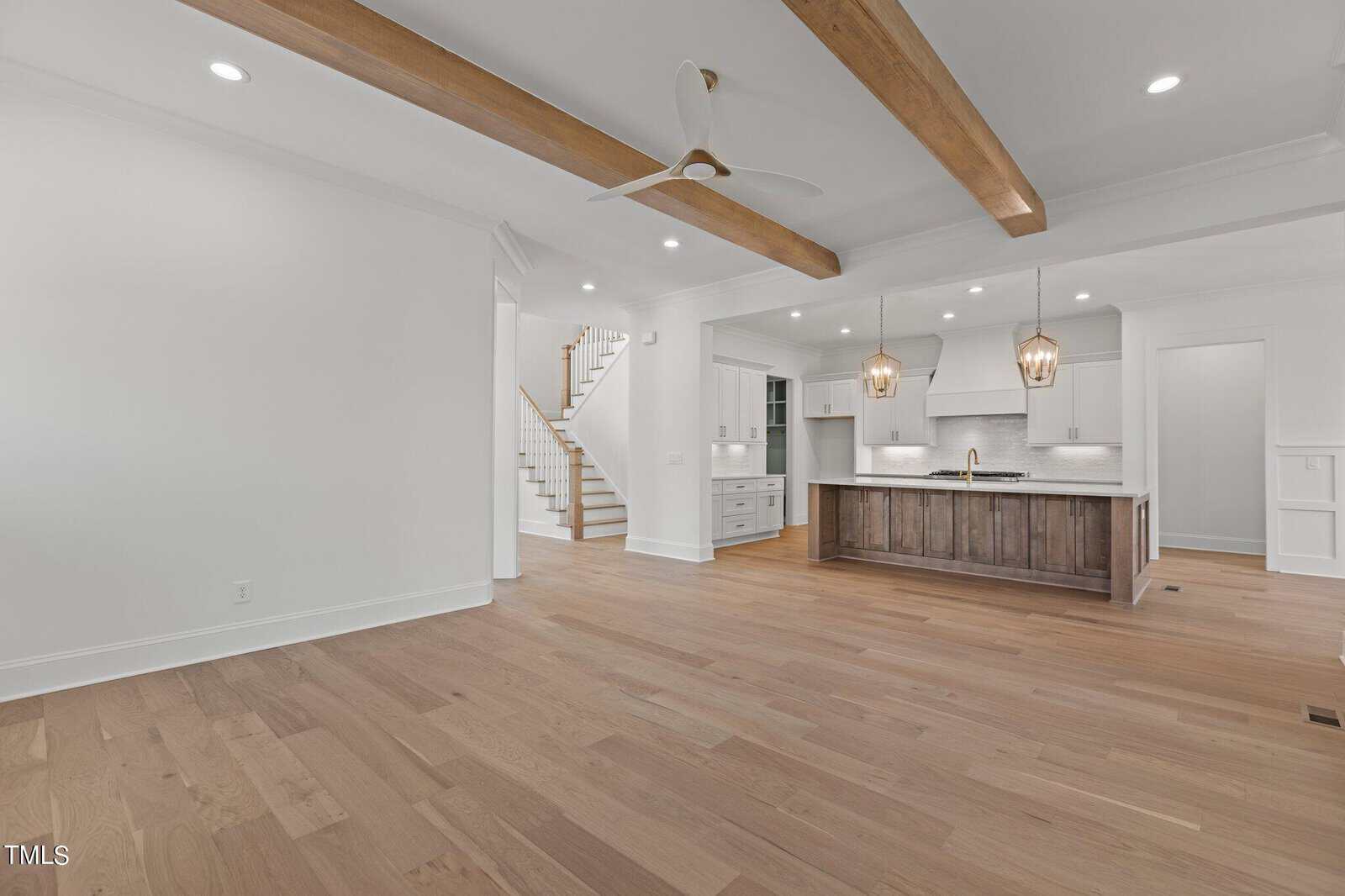 A spacious, empty room with wooden flooring features an open kitchen with a large island, pendant lights, and white cabinets. Exposed wooden beams accent the ceiling. A ceiling fan is installed, and a staircase is visible in the background.