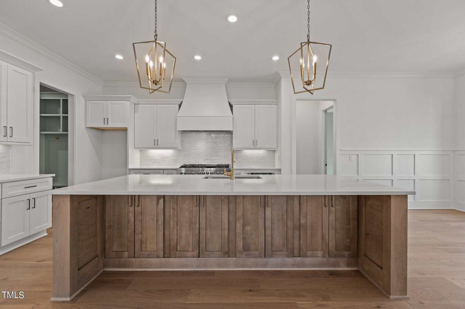 Modern kitchen interior featuring a large center island with wooden paneling and a white countertop. Two geometric pendant lights hang above. The background includes white cabinets, a stove with a hood, and light wood flooring.