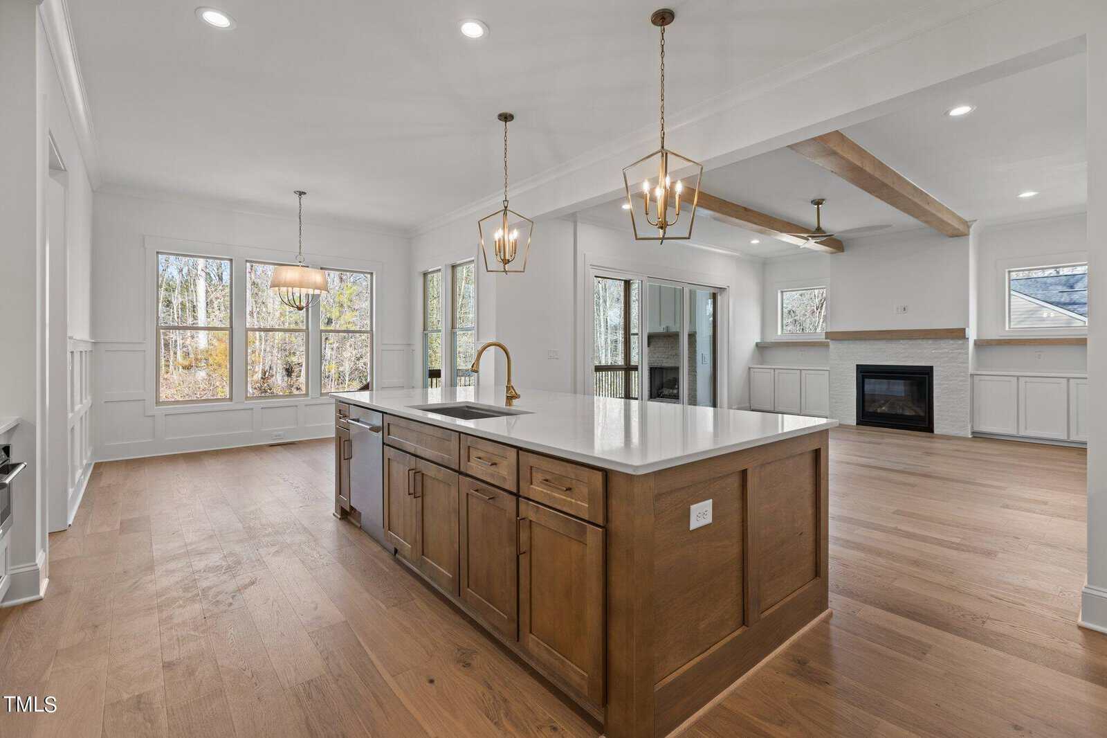 Modern open-concept kitchen and living area featuring a large central island with a white countertop and dark wood cabinetry. The space includes wooden floors, pendant lights, a fireplace, and large windows providing natural light.