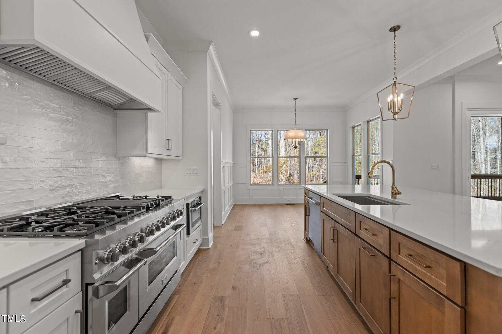 A spacious kitchen featuring a large gas stove and white cabinetry on the left, and a wooden island with a white countertop and sink on the right. Hanging light fixtures illuminate the area, while windows in the background provide natural light.