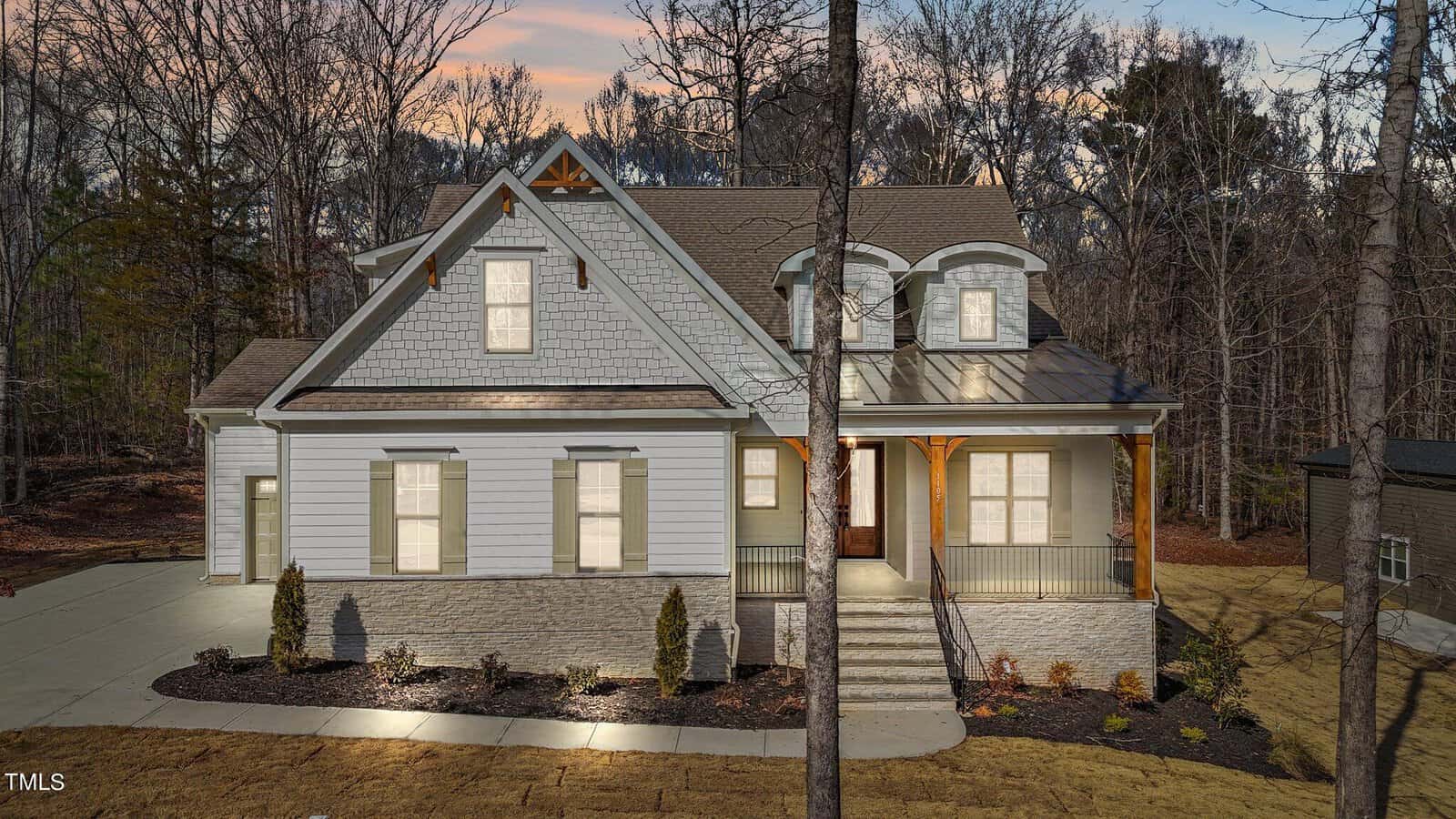 A charming two-story house at 1105 Springdale Drive, with a gray exterior, large windows, and a covered porch, sits amidst Wake Forest's wooded beauty. The driveway gently leads to the front steps, bordered by small shrubs at the base.