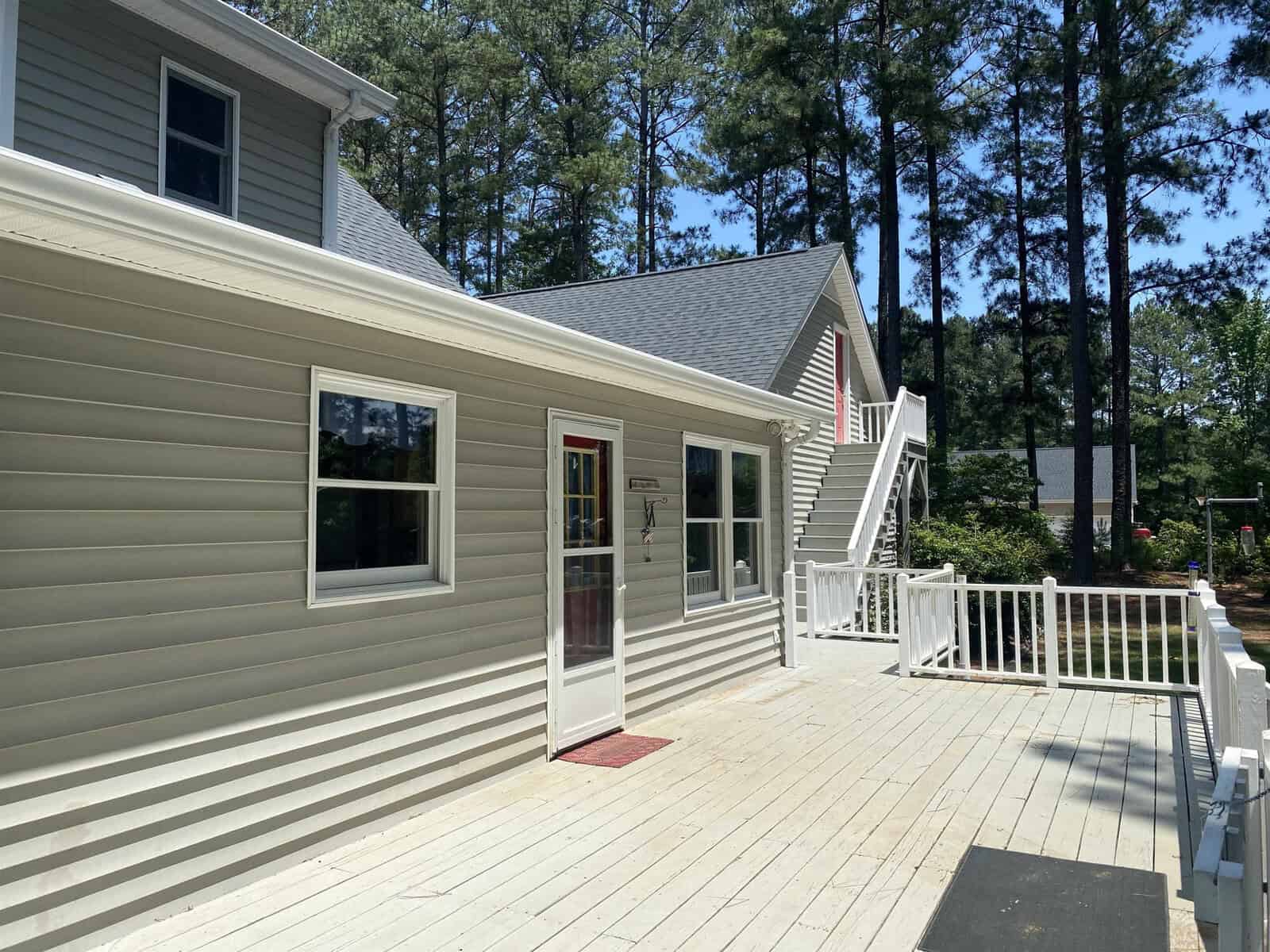 A sunlit house with light gray siding features a spacious deck and a stairway leading to the upper level. Surrounded by tall pine trees, the scene conveys a peaceful, wooded setting.