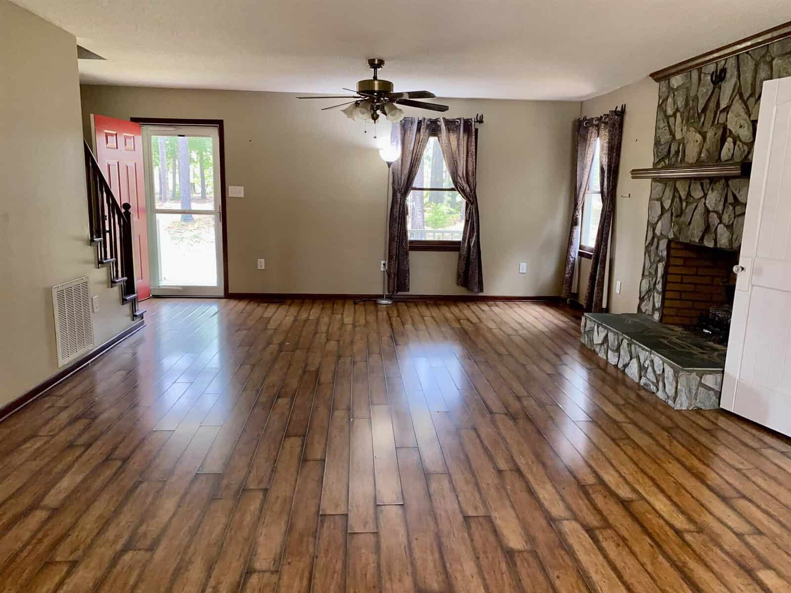 Spacious living room with wooden floors, large windows with dark curtains, a ceiling fan, and a stone fireplace. An open entrance door reveals a view outside, and a staircase is visible to the left.
