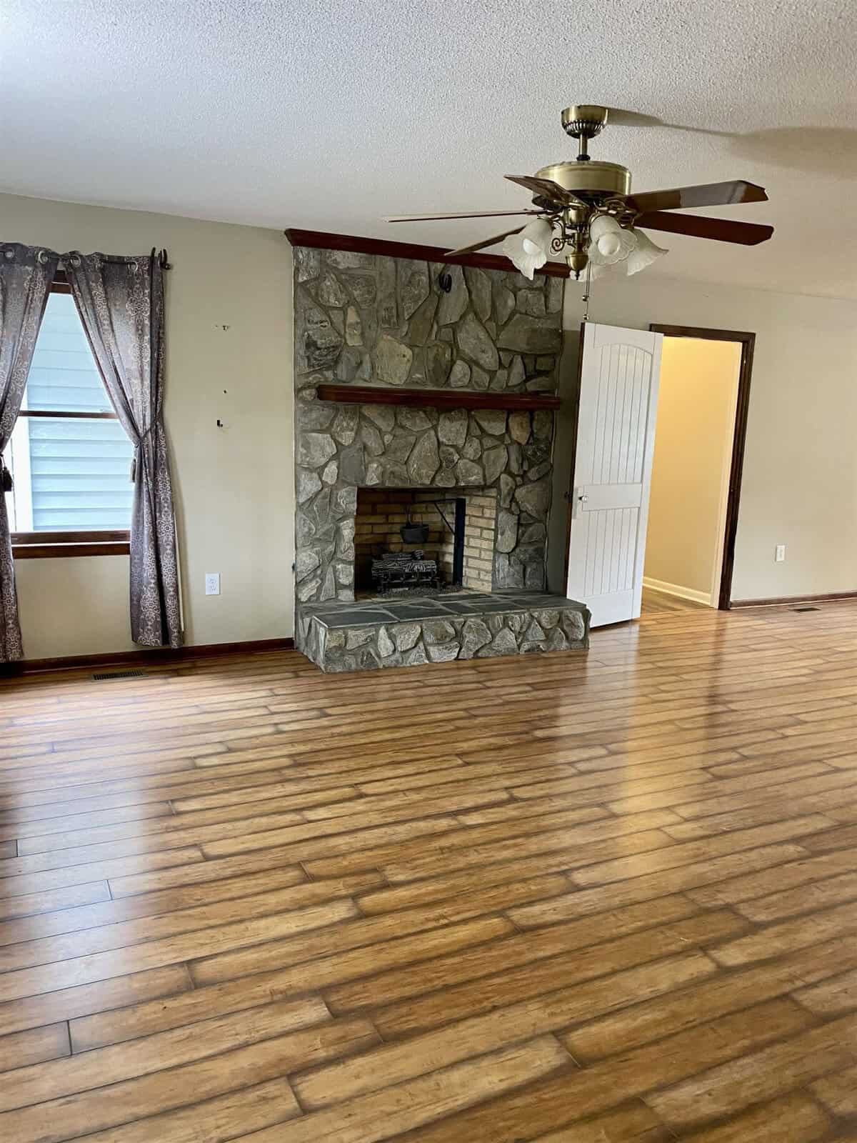 A spacious living room with wooden floors and a stone fireplace. The room includes a ceiling fan and a large window with patterned curtains. An open door leads to another room, adding to the airy feel of the space.