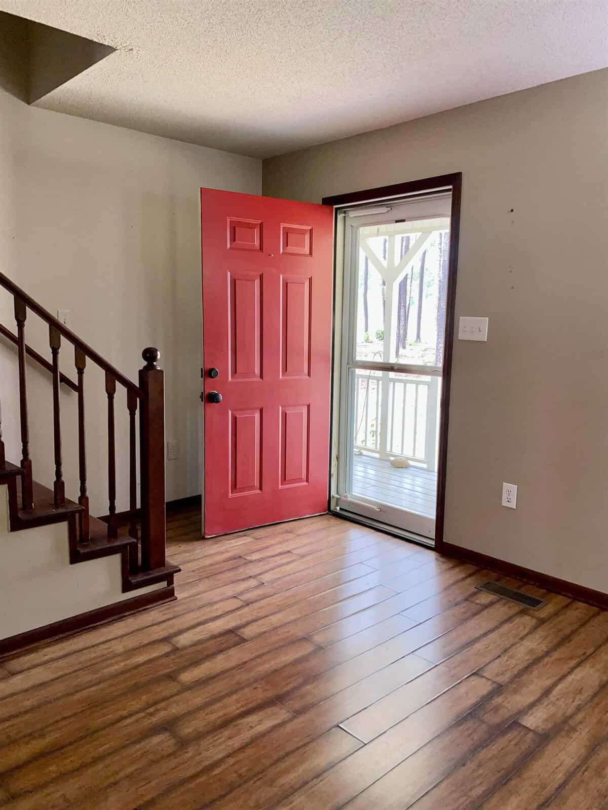 A room with hardwood flooring and a red door partially open, revealing a glass storm door. Wooden stairs with a dark railing are on the left, and the wall is painted light gray. Sunlight streams in through the doors, illuminating the space.