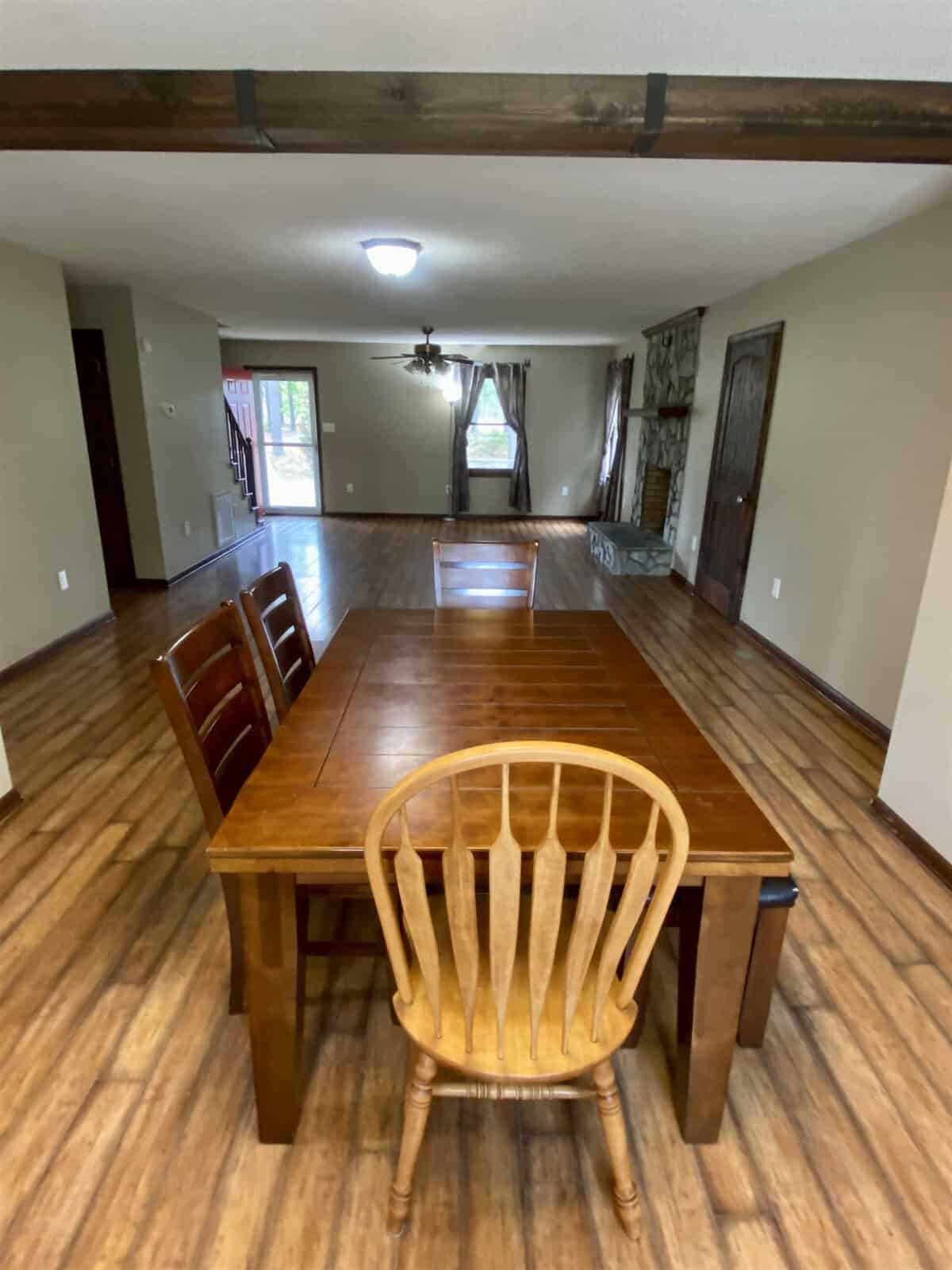 A spacious dining room with a wooden table and chairs on a hardwood floor. The room features light-colored walls, a ceiling fan, and a fireplace in the background. Large windows provide natural light.