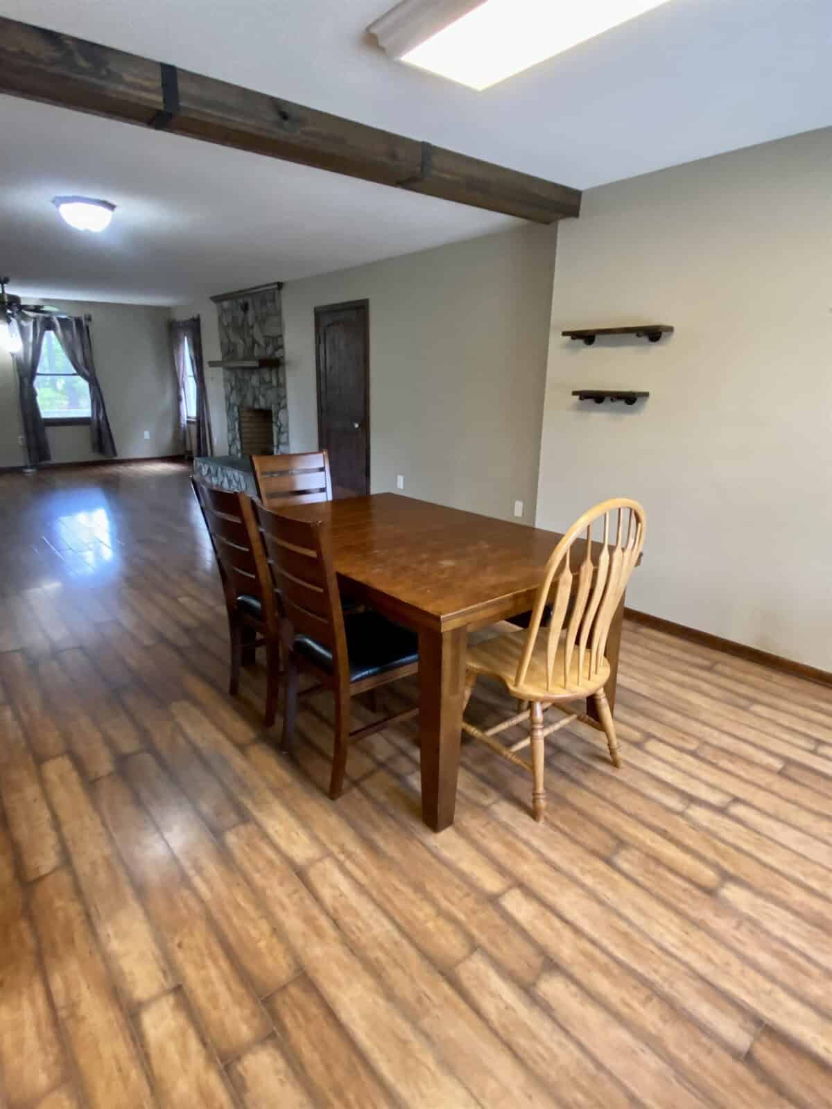 A dining area with a wooden table and four chairs on a hardwood floor. The room has beige walls, two small shelves, and a view of the adjacent living area with windows and curtains in the background.