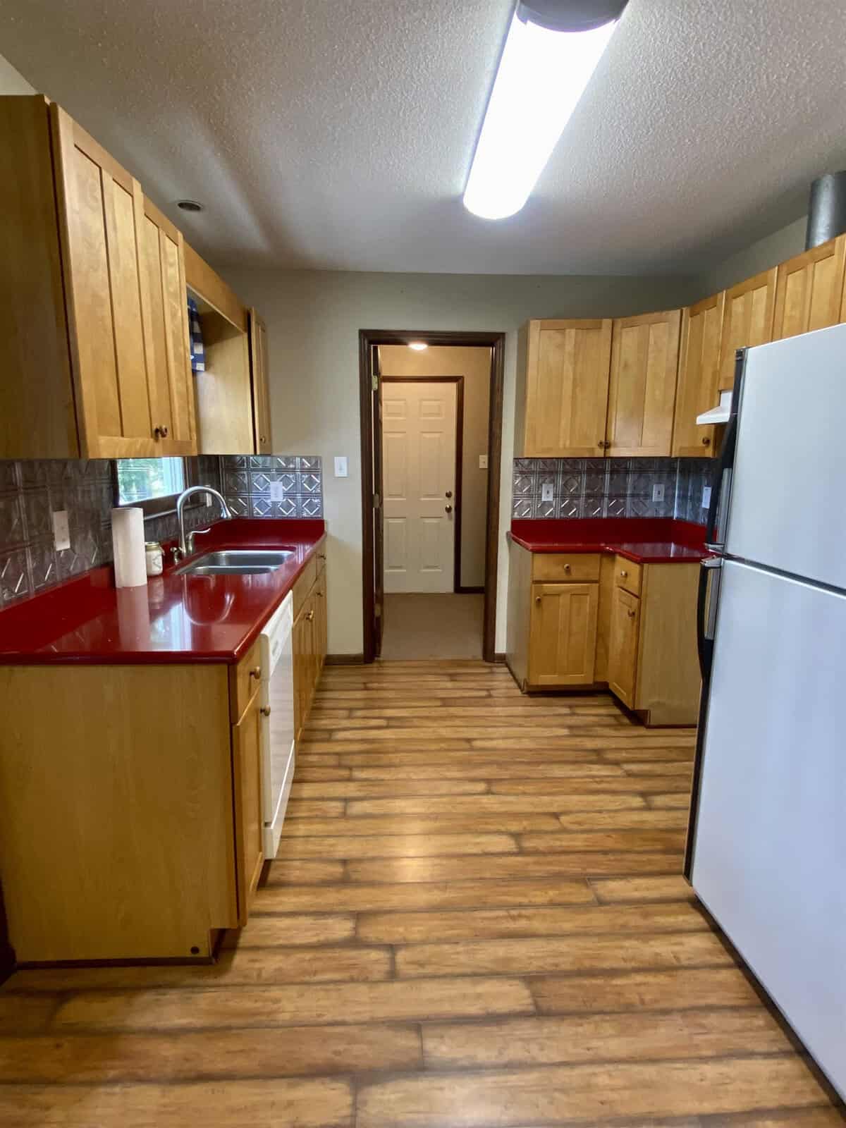 A kitchen with wooden cabinets and red countertops. It features a white refrigerator, a dishwasher, wood-patterned flooring, and a window above the sink. The room is well-lit by an overhead fluorescent light. A door is visible in the background.