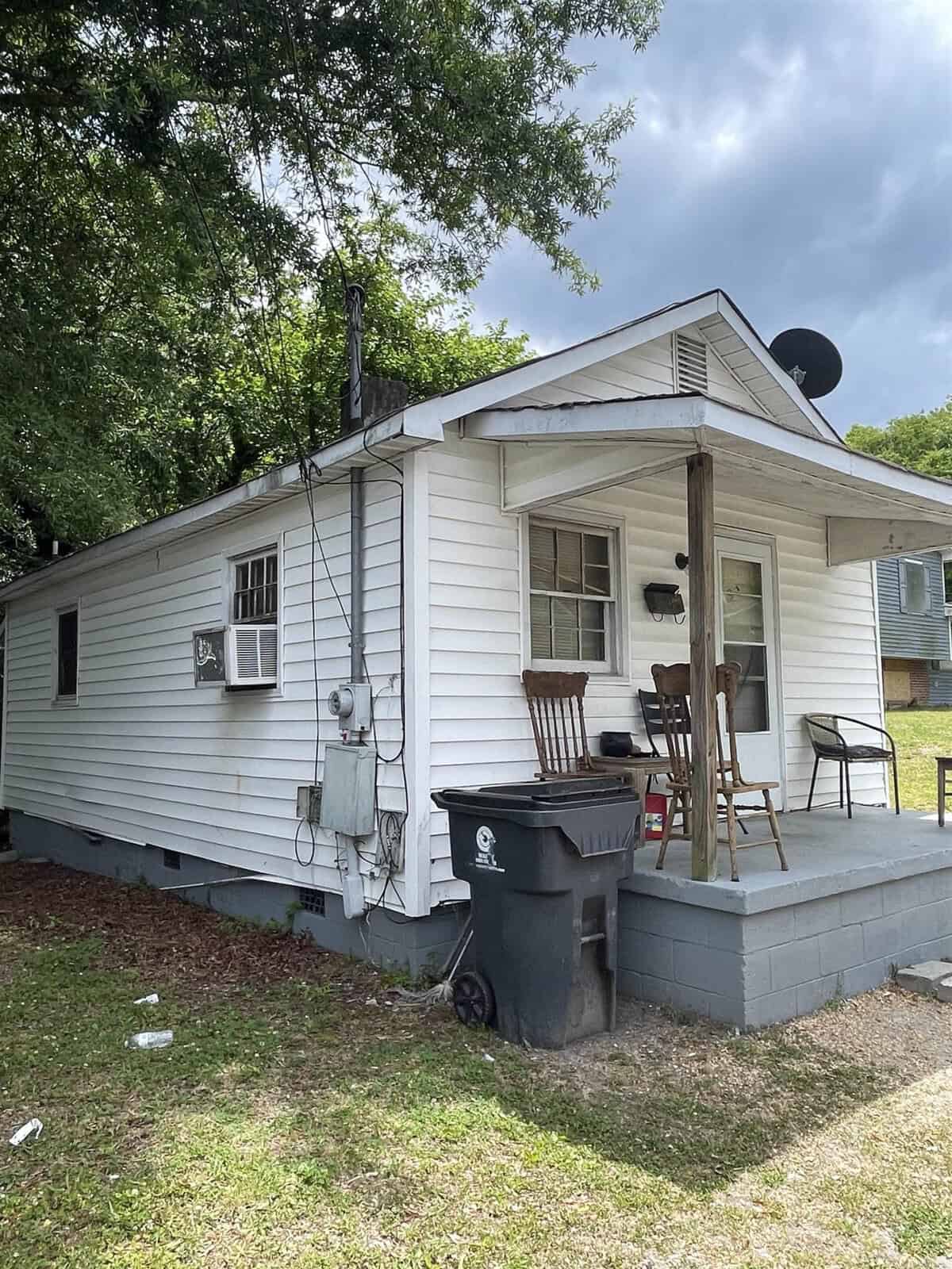 A modest, single-story white house with a front porch, outfitted with two chairs, and a satellite dish on the roof. a trash bin and miscellaneous items are scattered in the yard.