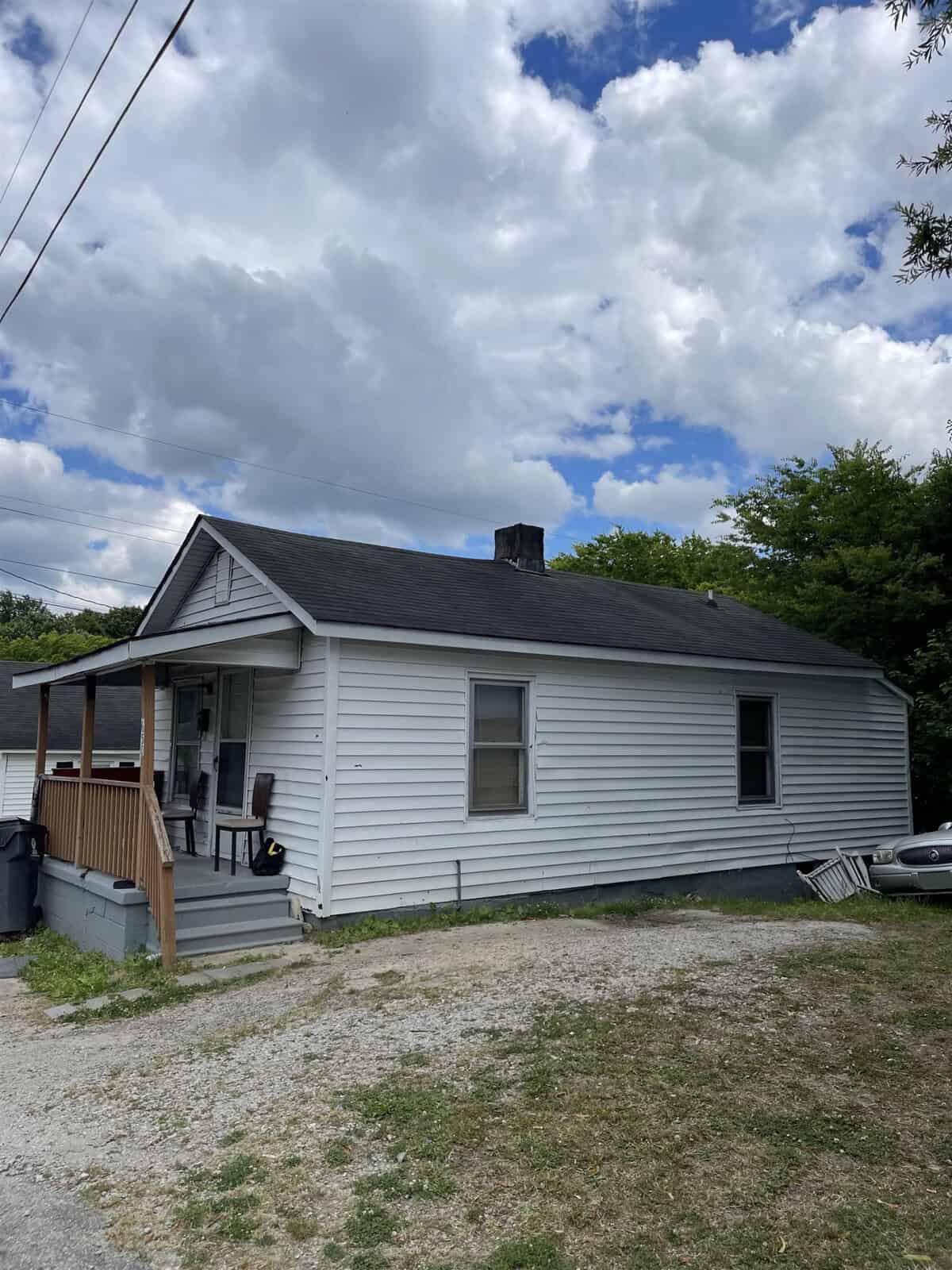 A small, single-story house with white siding, featuring a front porch with a wooden railing, is set against a partly cloudy sky. there is a gravel driveway on the side with a car parked, and utility lines visible overhead.