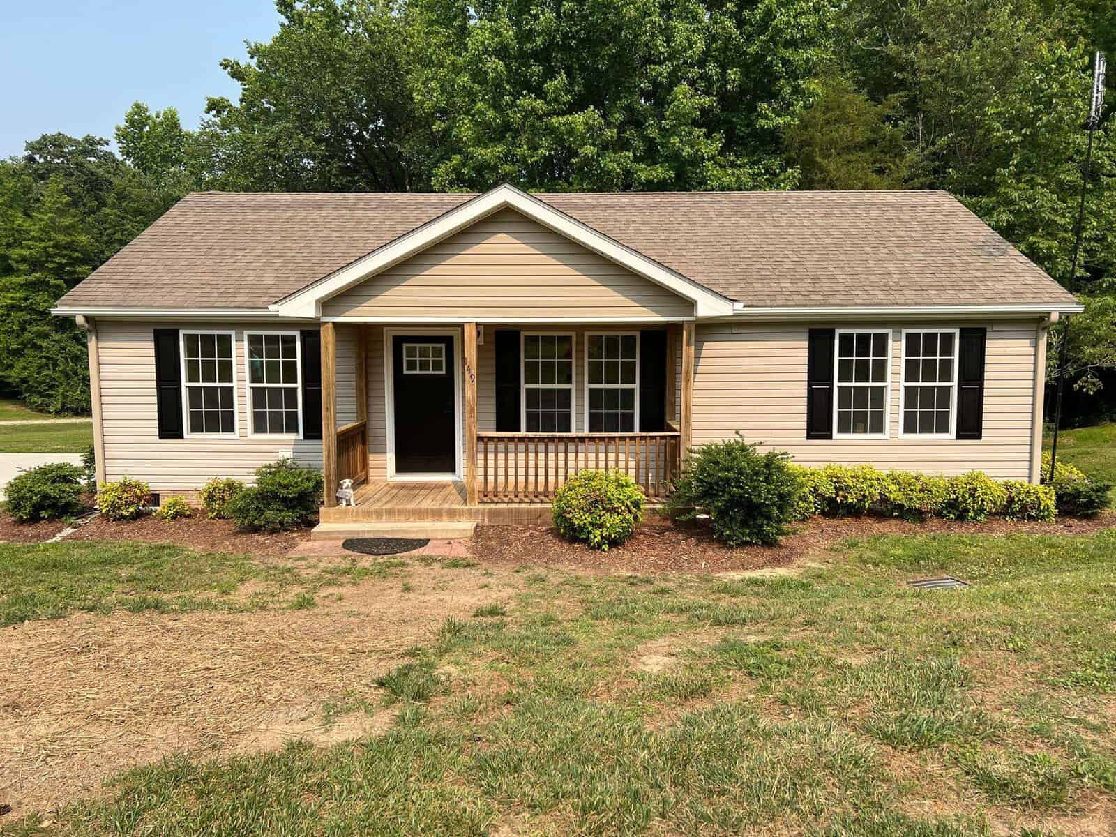 A single-story beige house at 149 Magnolia Court features a peaked roof, black shutters, and a small front porch. It's surrounded by a green lawn, bushes, and trees in the background reminiscent of Manson's serene charm. A pathway gracefully leads to the front door.